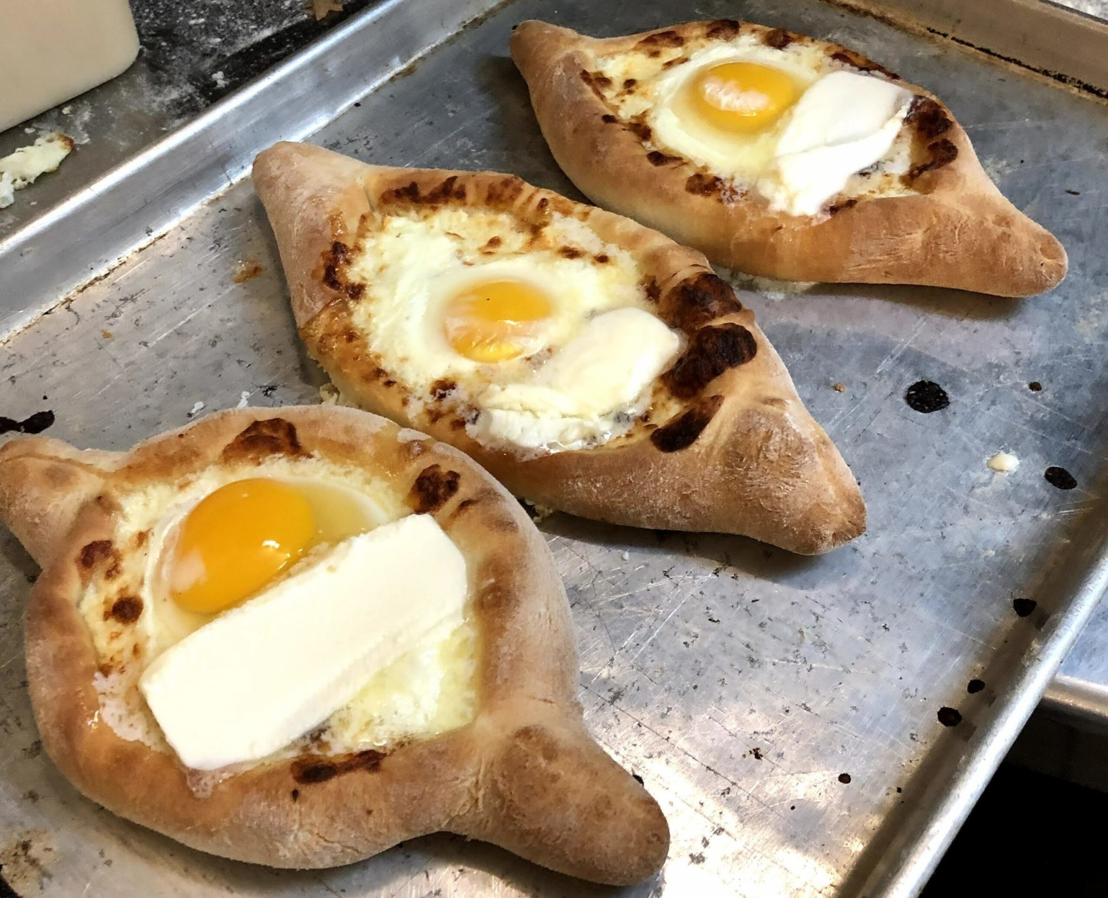 Three boat-shaped breads topped with cooked eggs and a slice of white cheese or butter sit on a metal baking tray.
