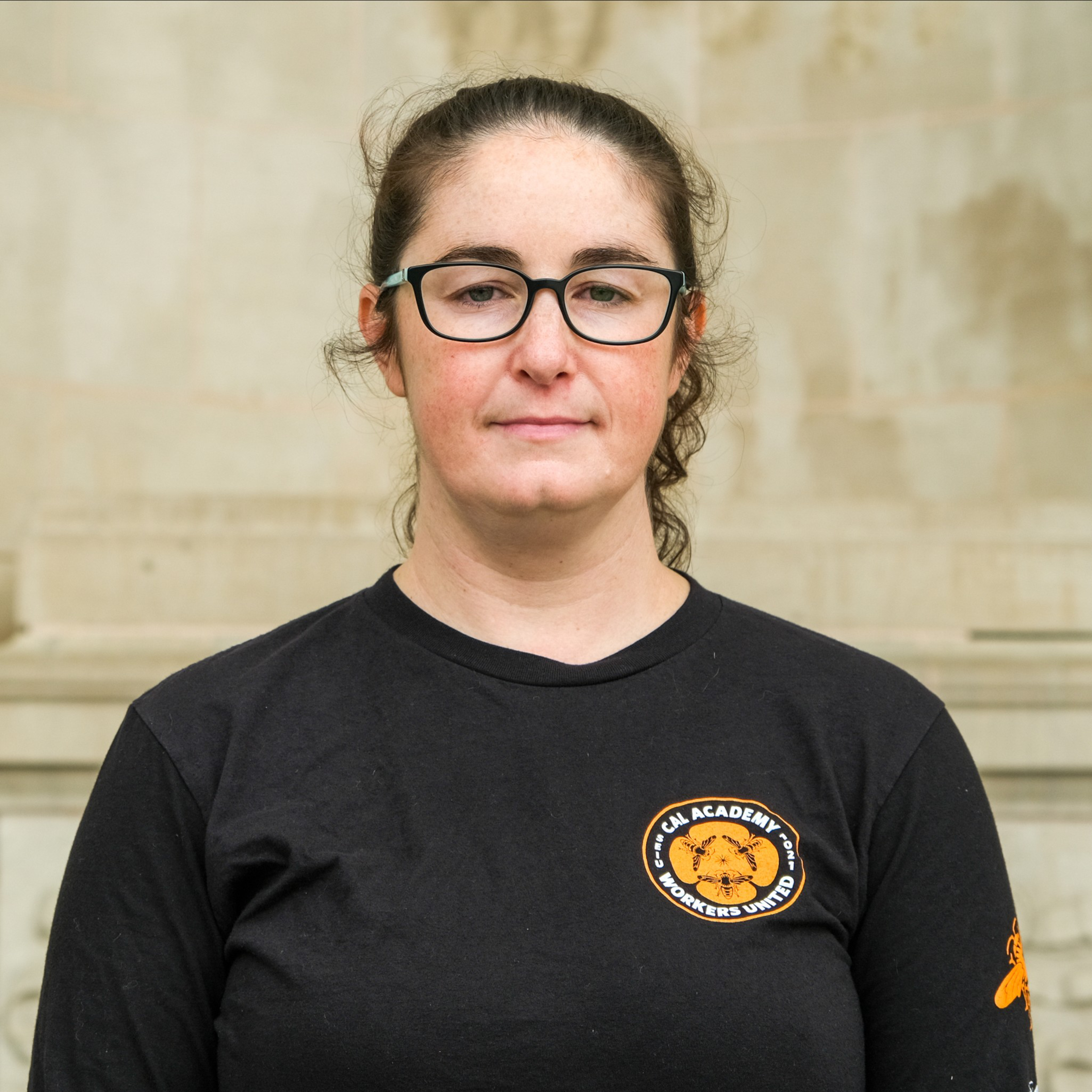 A woman with glasses and a ponytail, wearing a black T-shirt with a logo &quot;Cal Academy Workers United&quot;, stands against a beige wall.