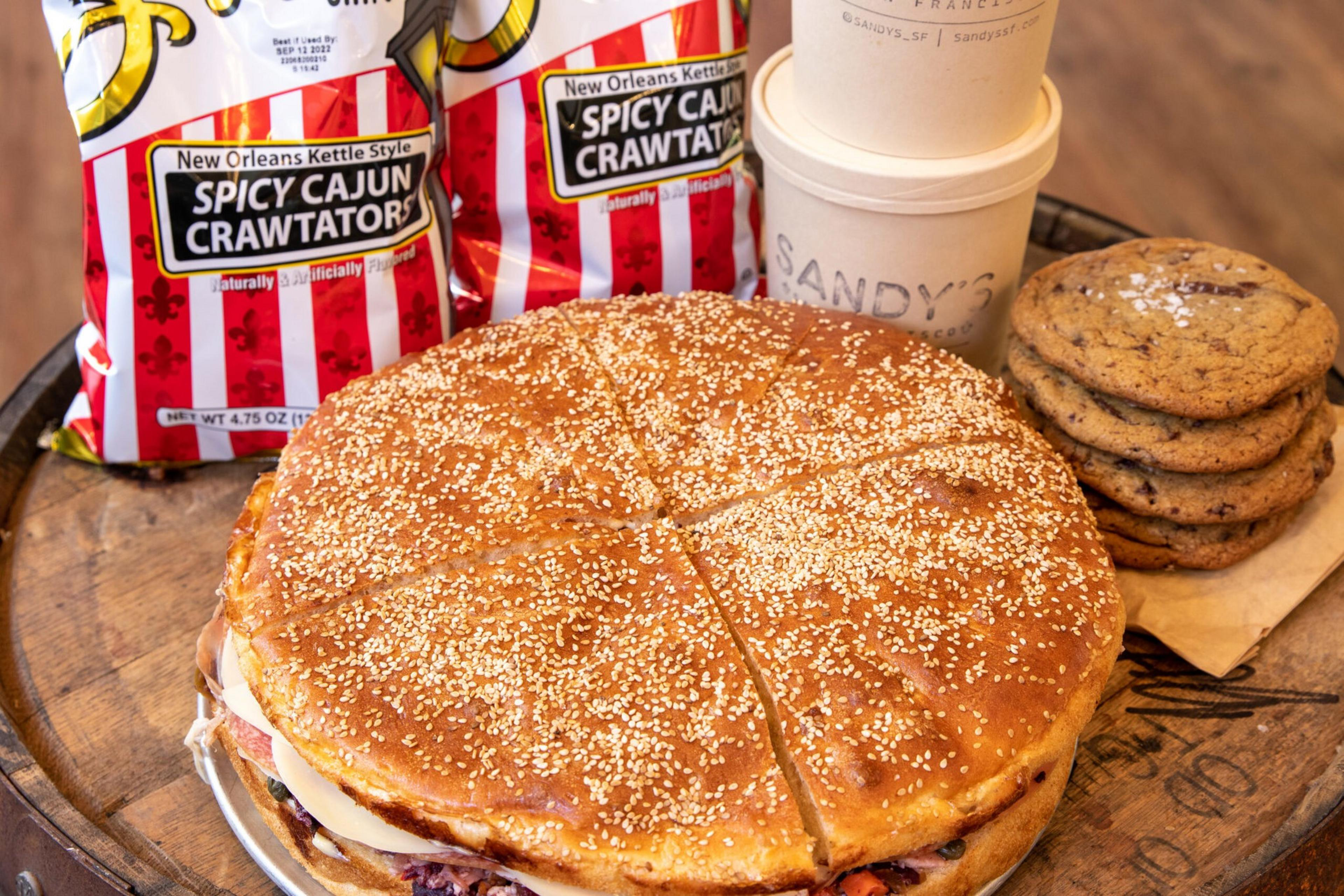 A large sesame seed sandwich sits on a wooden surface, surrounded by two bags of Spicy Cajun Crawtators, a stack of chocolate chip cookies, and a Sandy's cup.
