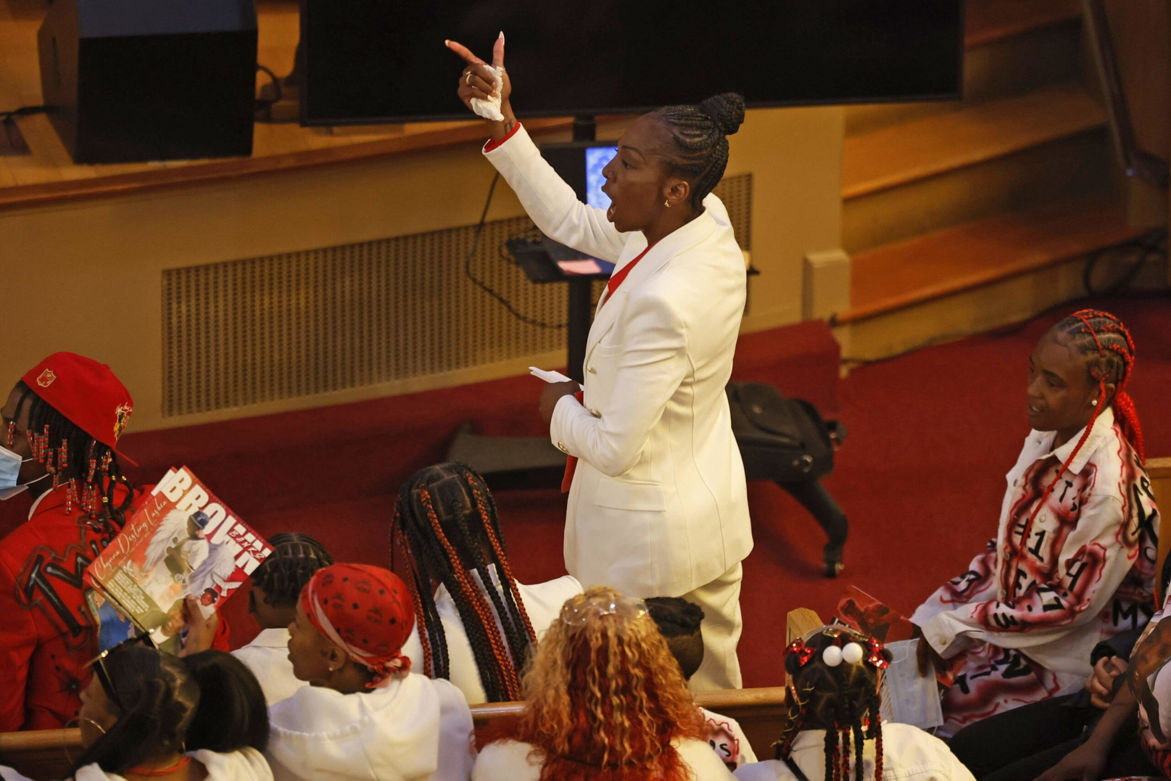 A woman in a white suit speaks passionately to a seated audience, with one person holding a colorful magazine and others wearing vibrant hairstyles and clothing.