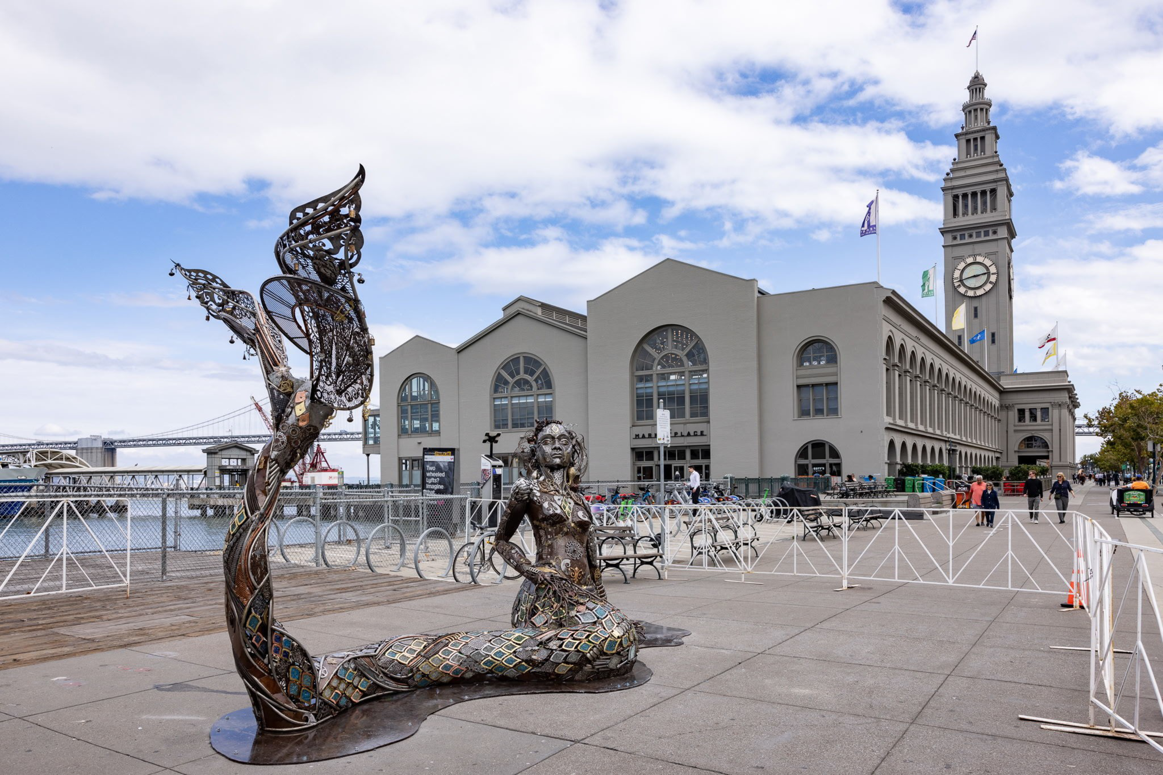 A large metal mermaid sculpture sits on a waterfront plaza with a clock tower and arched building in the background under a partly cloudy sky.