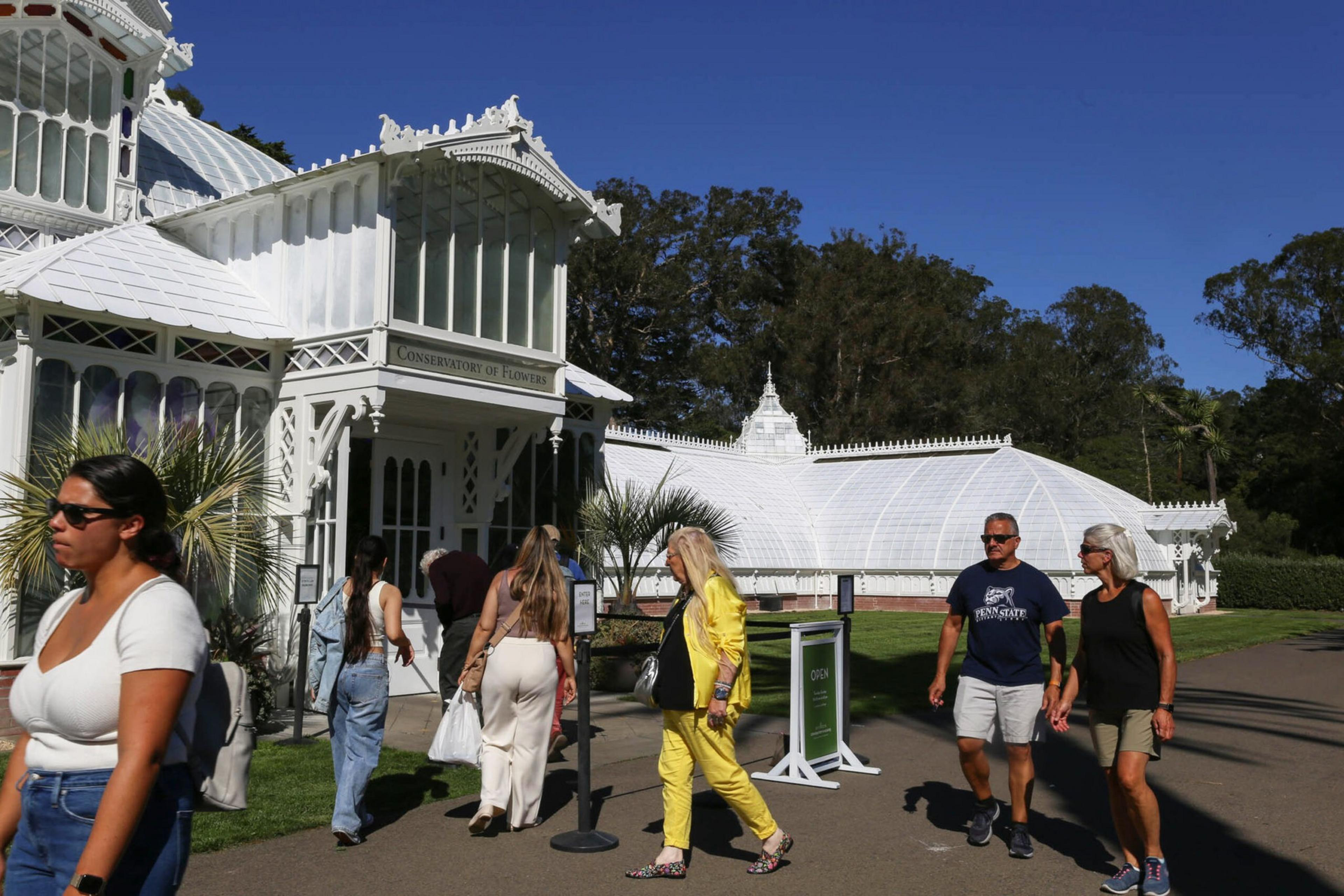 Visitors walk near a large white conservatory building labeled "Conservatory of Flowers" under a clear blue sky.