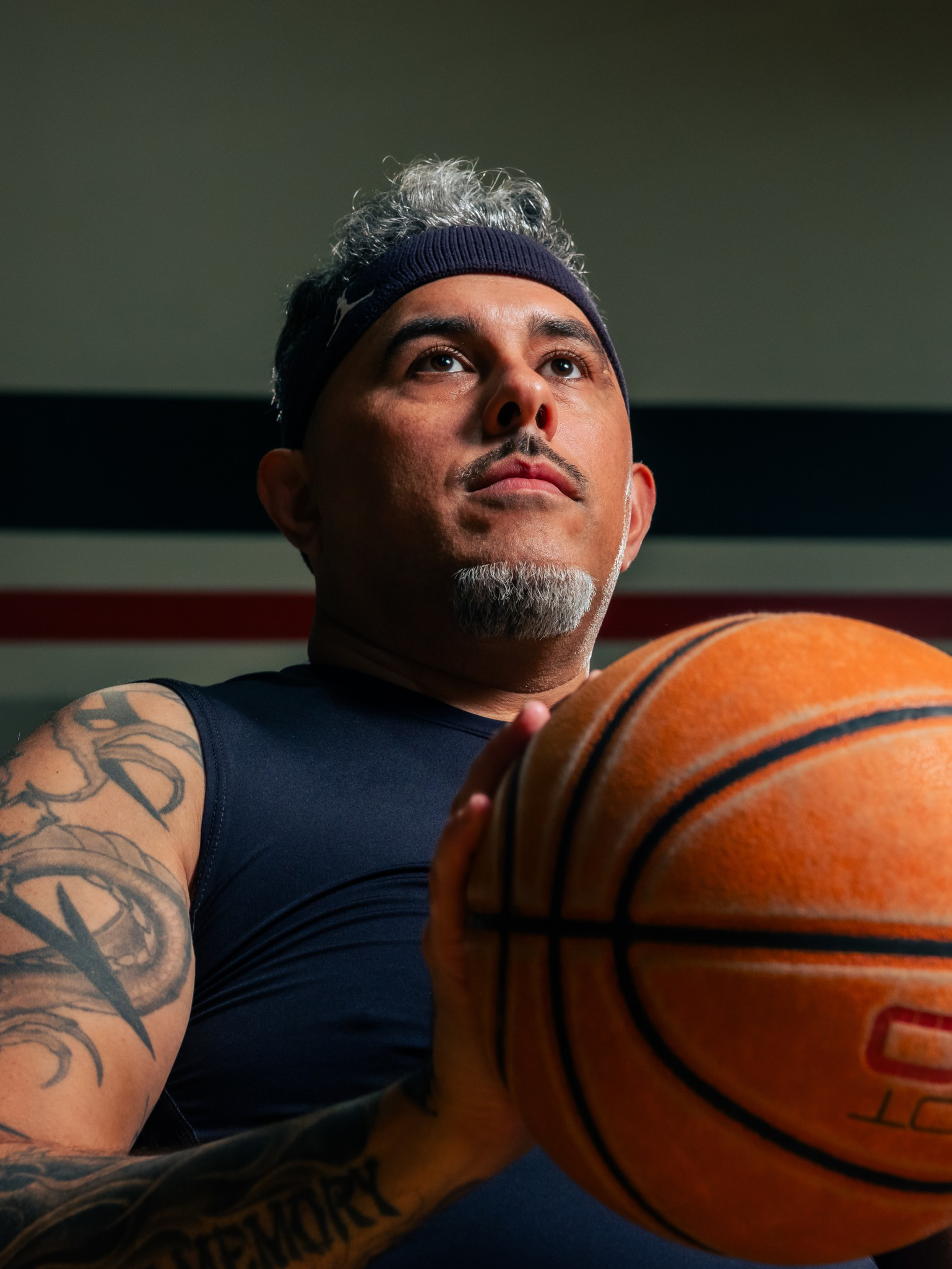 A man with gray hair, a goatee, a headband, and tattoos is holding a basketball, looking focused and ready to shoot or pass.