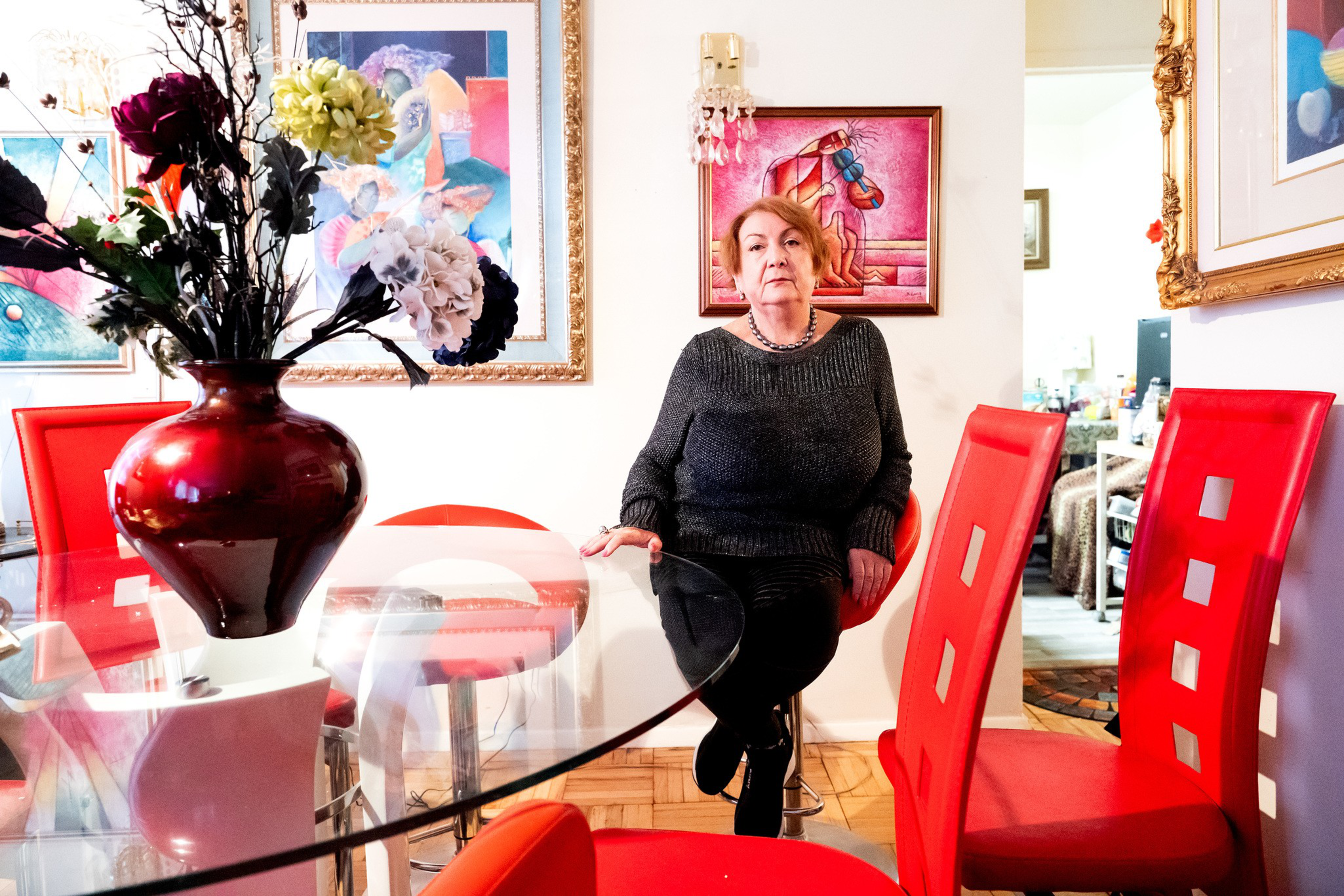 A woman in a dark sweater sits on a chair in a dining room with red chairs, a glass table, vibrant floral centerpiece, and colorful framed paintings.