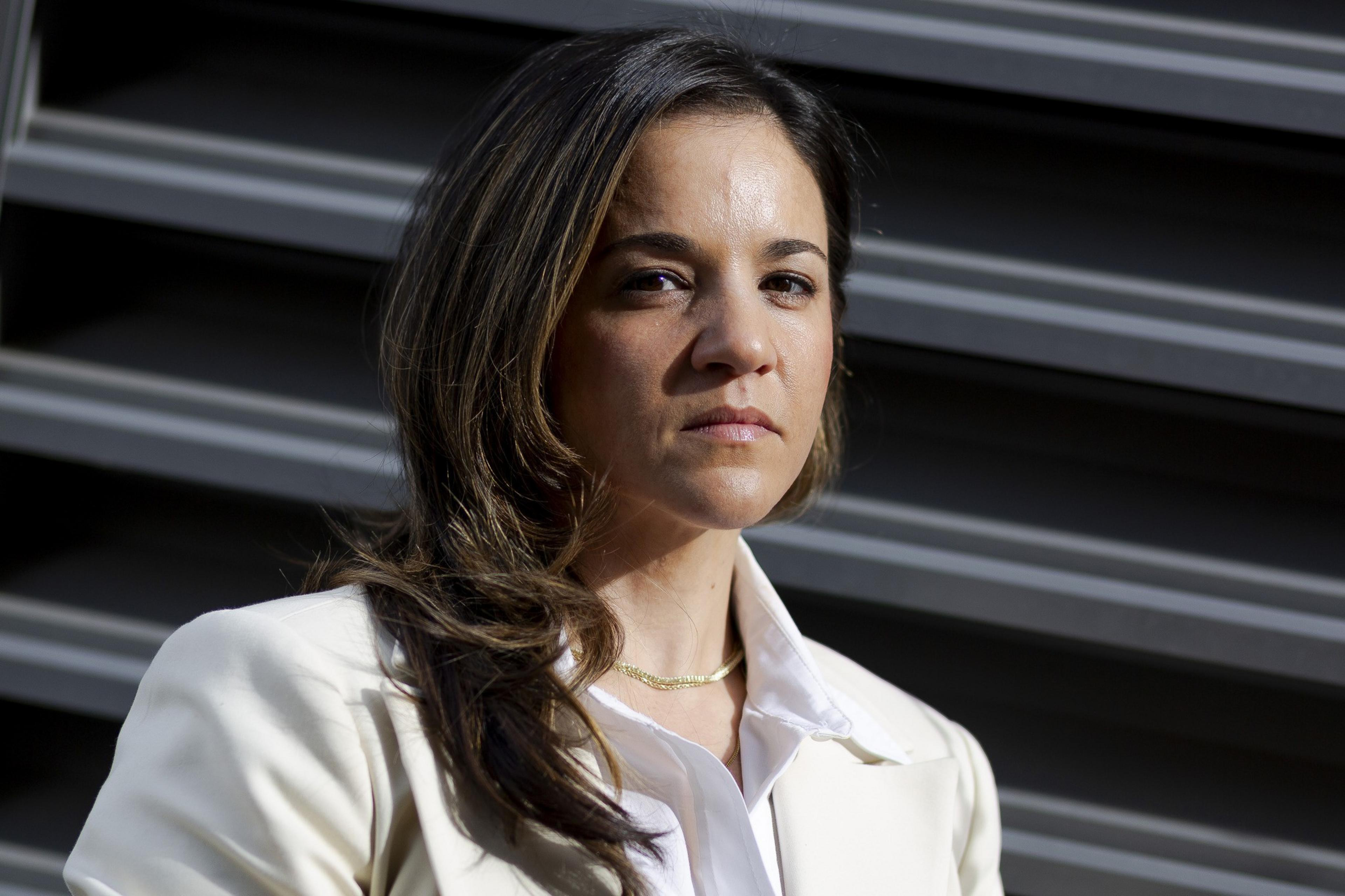 A woman with long, dark hair stands against a backdrop of horizontal metal slats. She wears a light-colored blazer and a serious expression.