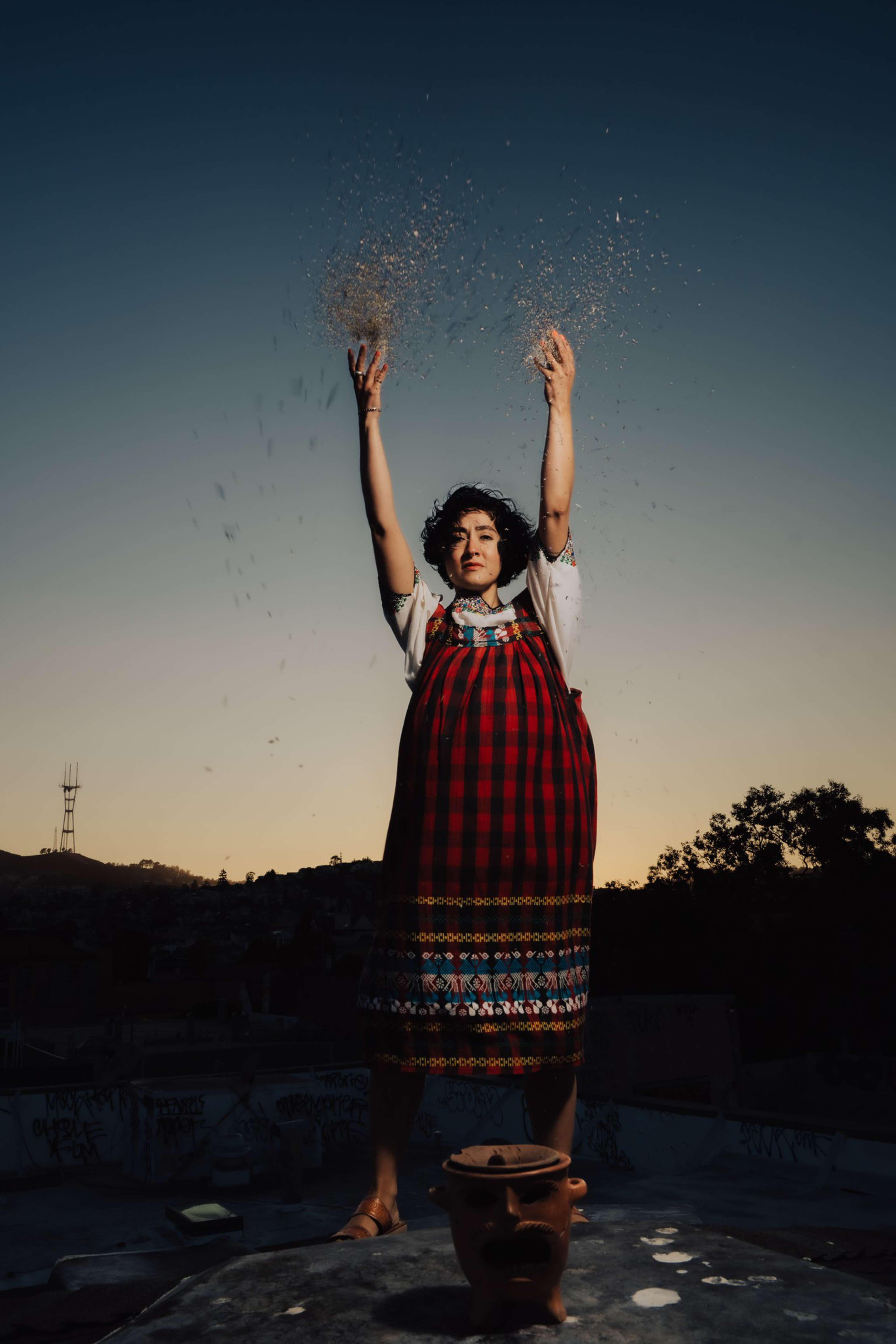 A woman in a colorful dress stands on a rooftop at sunset, raising her arms while scattering glitter or small particles into the air.