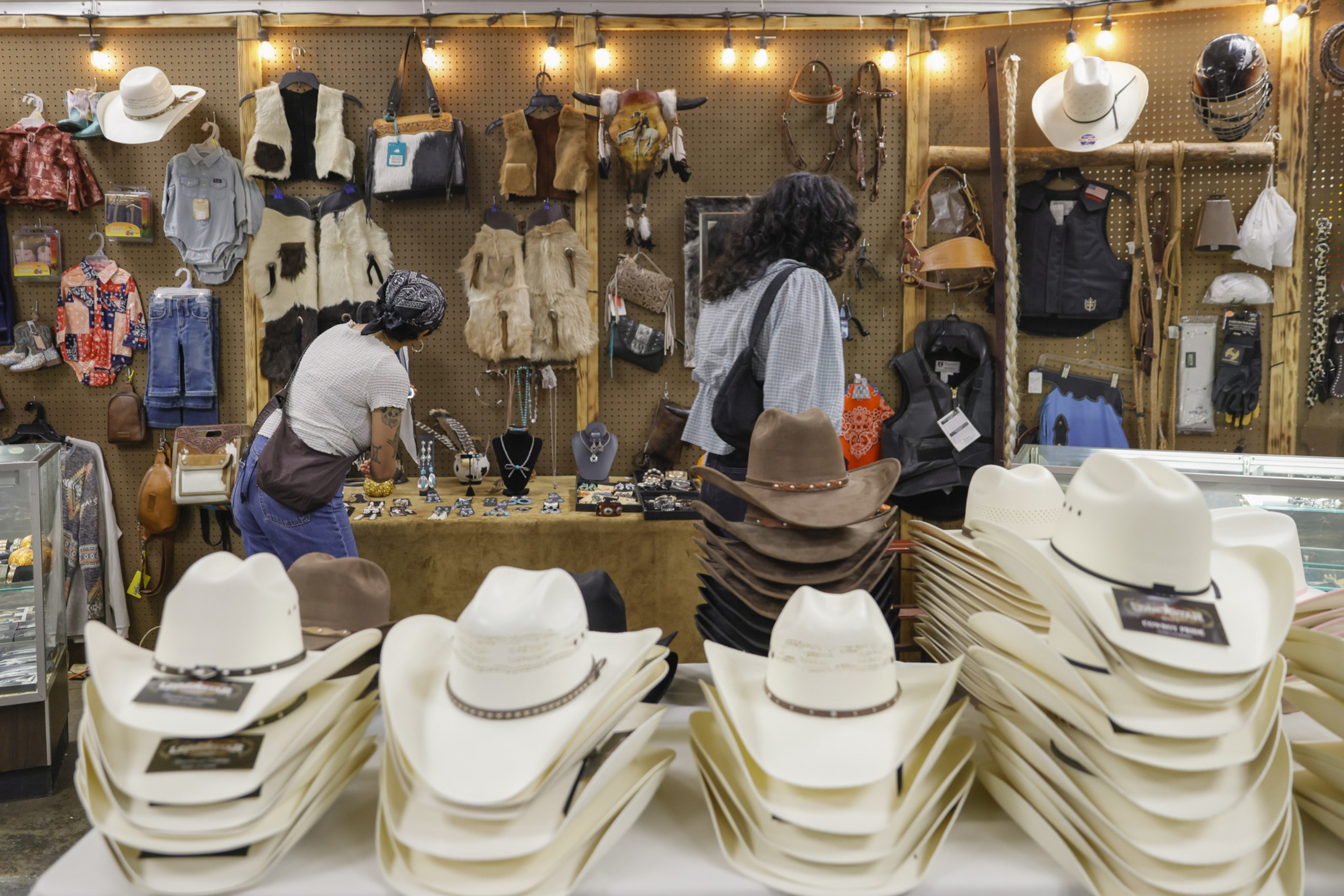 Stacks of white and brown cowboy hats sit on tables, with leather vests, bags, jewelry, and other western accessories displayed on walls and shelves.