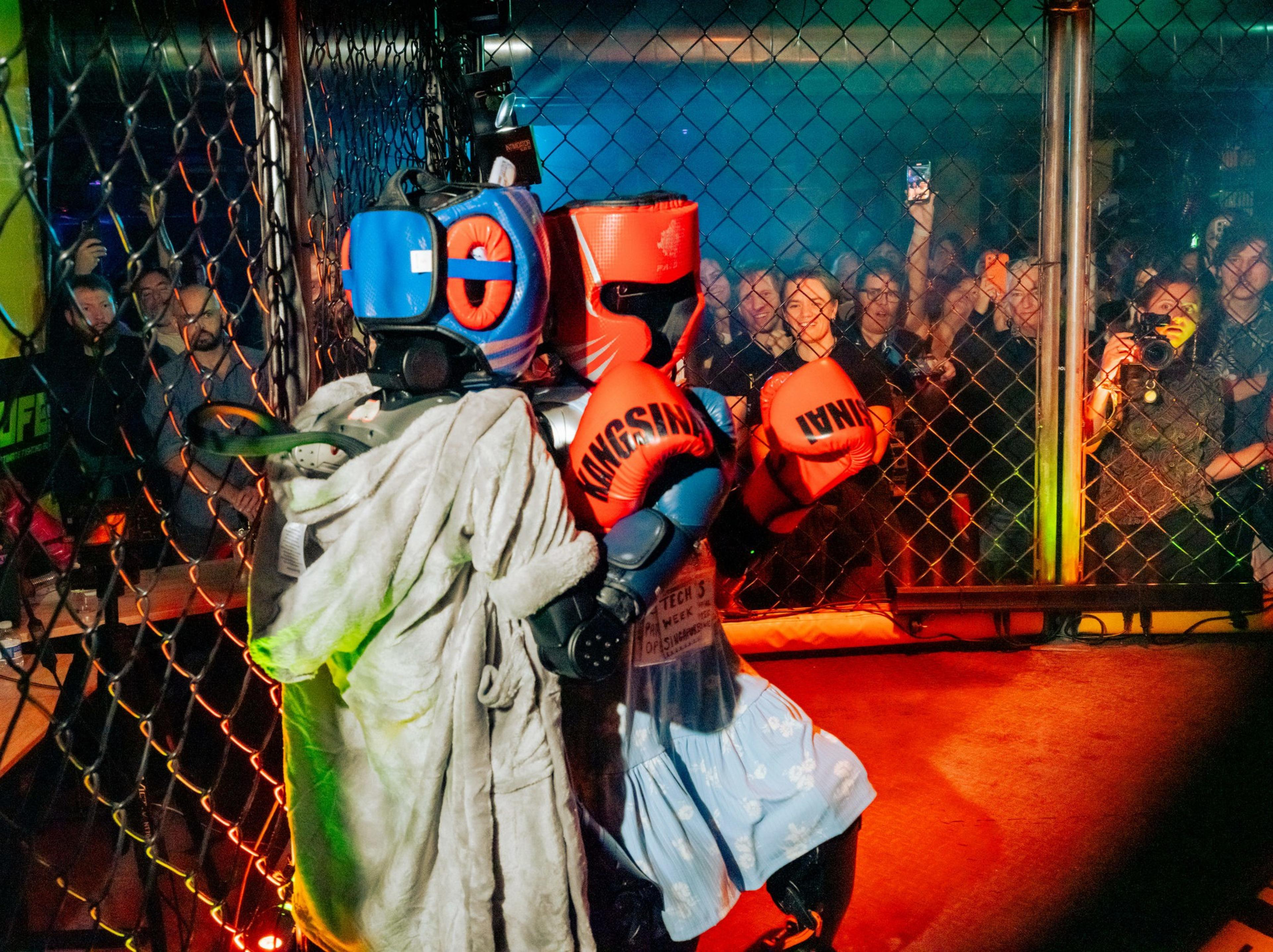 Two robots wearing boxing gloves and protective headgear face each other inside a metal cage ring, surrounded by an audience watching intently.