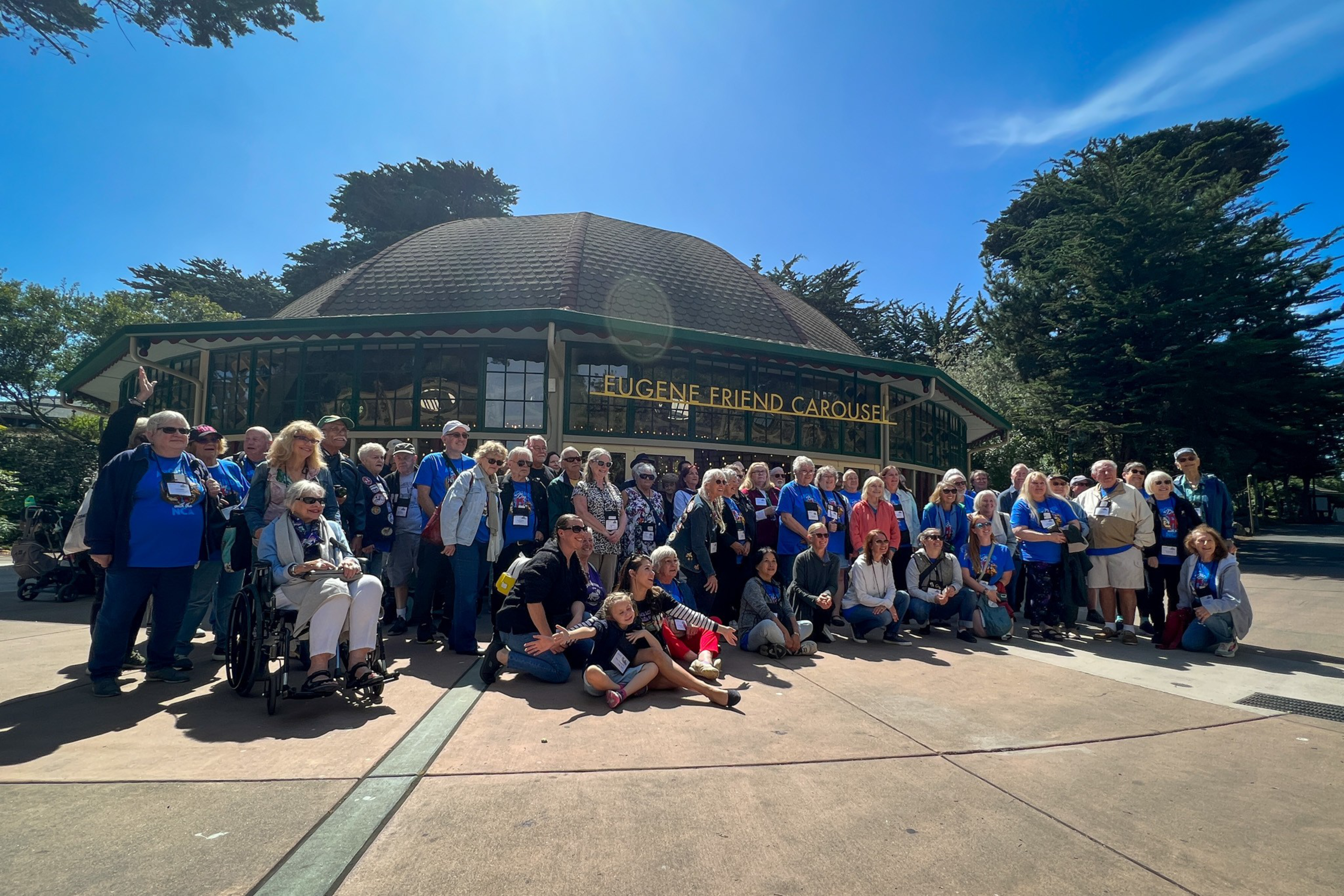 A large group of people of various ages pose together outside a round building labeled "Eugene Friend Carousel" under clear blue skies.