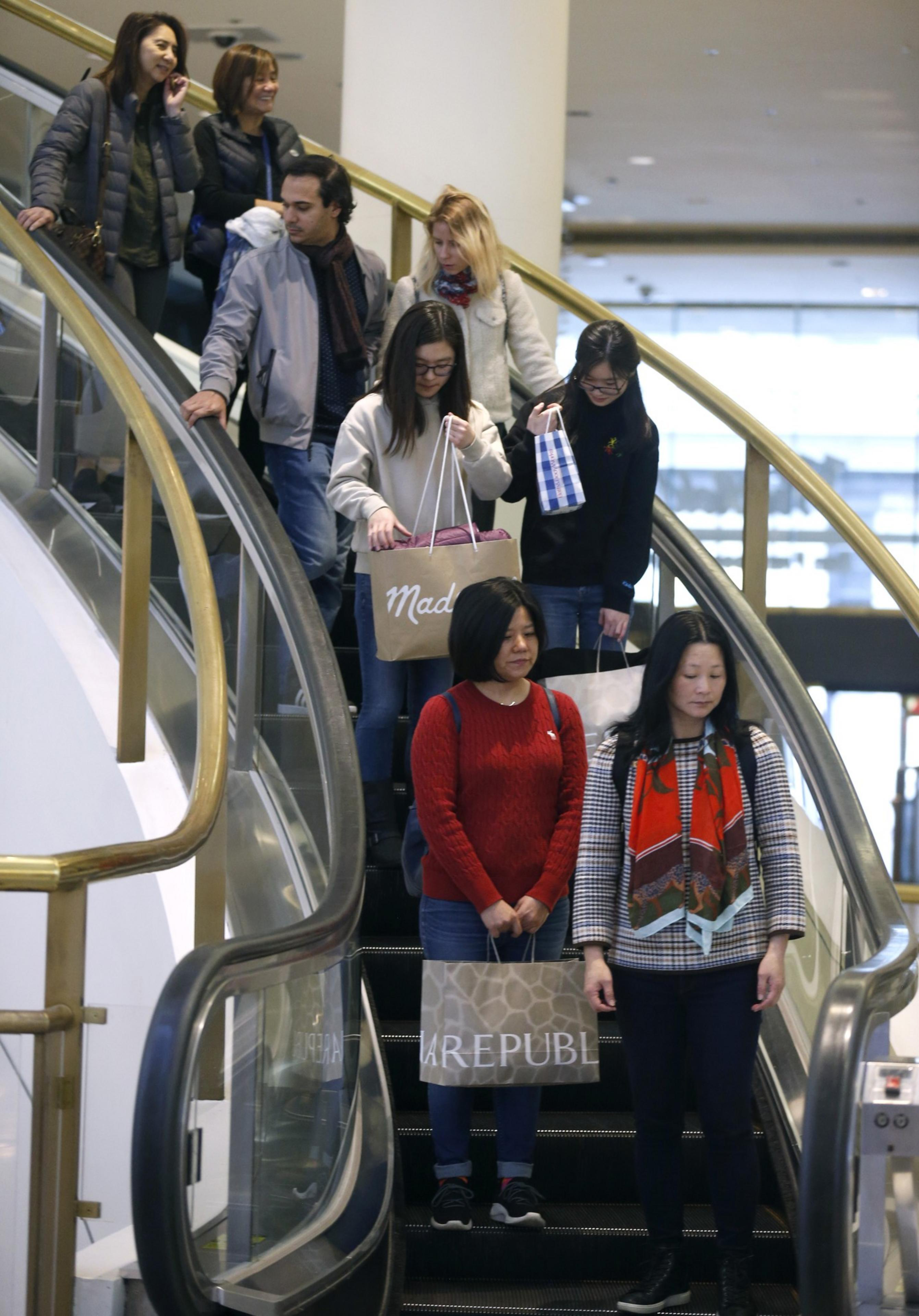People are descending on an escalator, holding shopping bags. There's a mix of ages and styles.