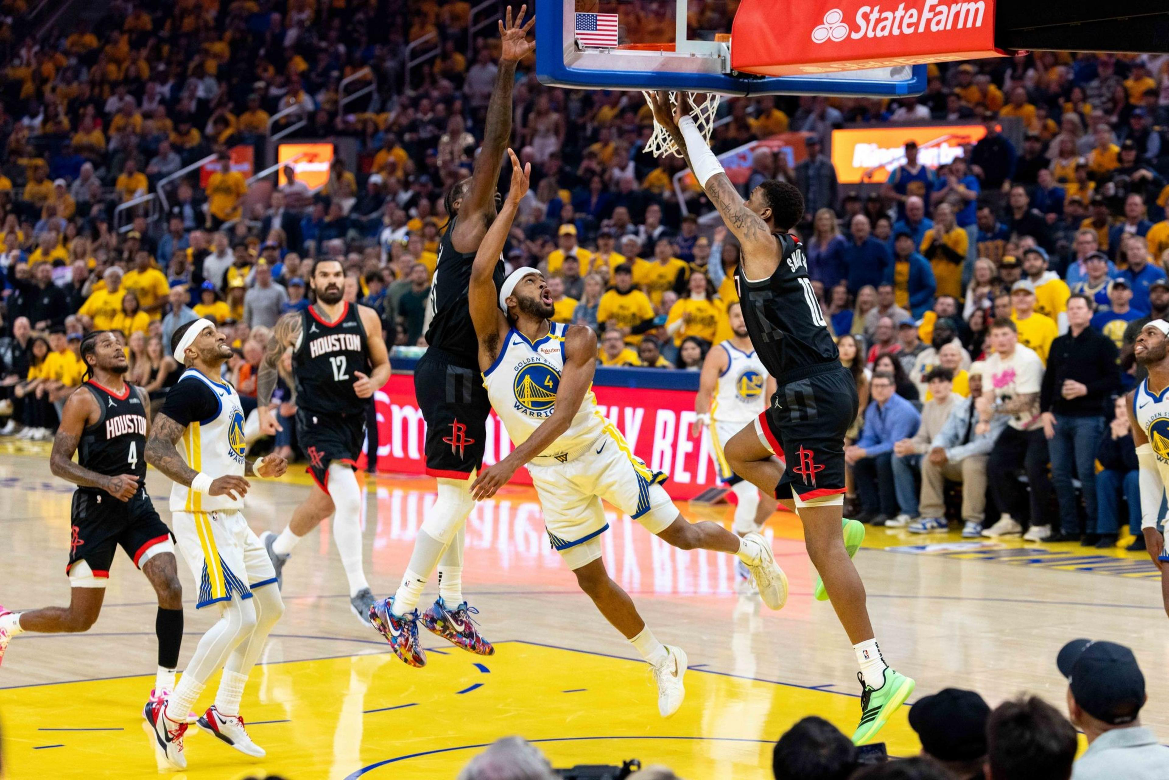 In a basketball game, a player in a black uniform jumps to make a layup while being defended by players in white and blue. The audience watches intently from the stands.