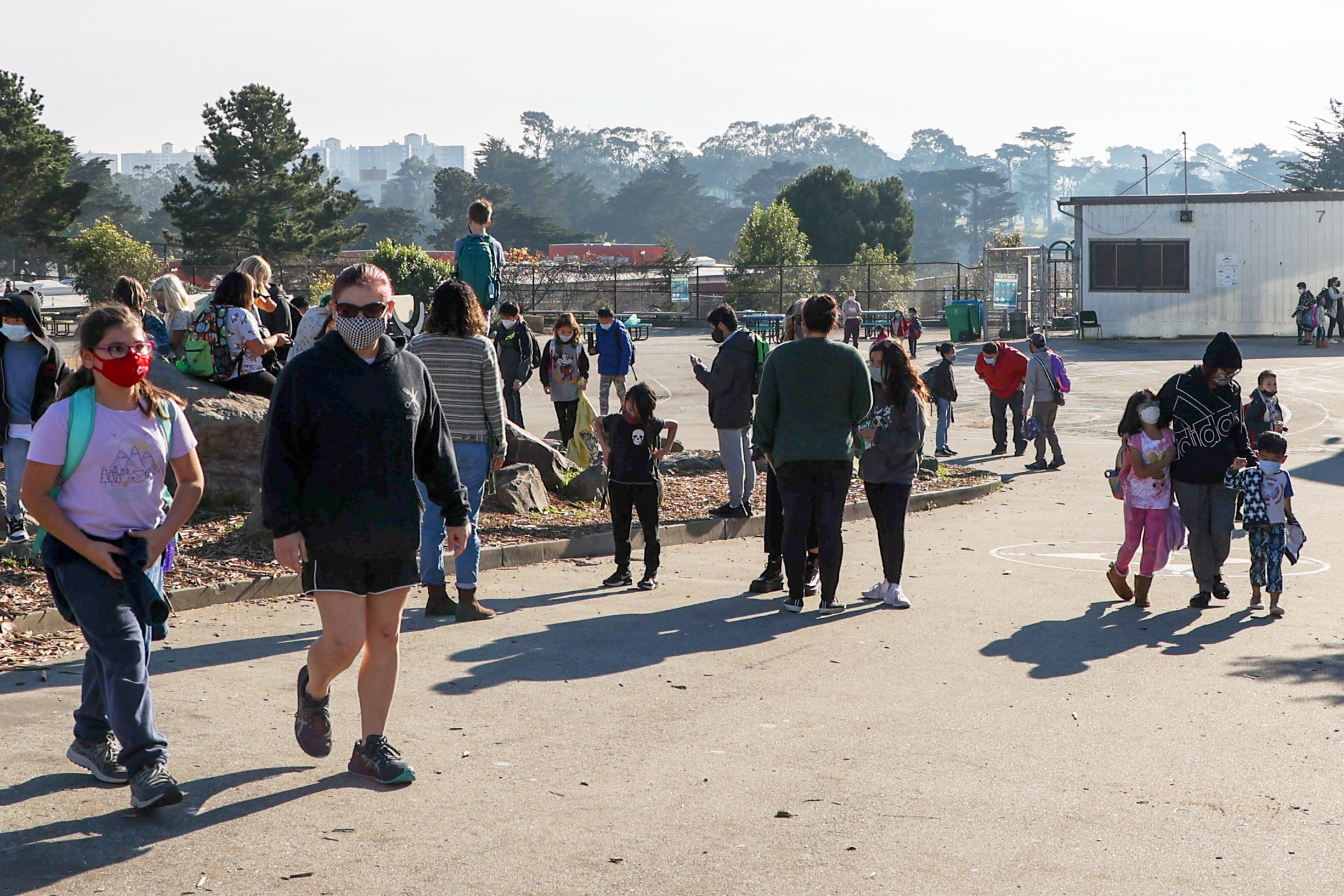 Children and adults wearing masks walk and gather outside on a sunny schoolyard with trees and buildings in the background.
