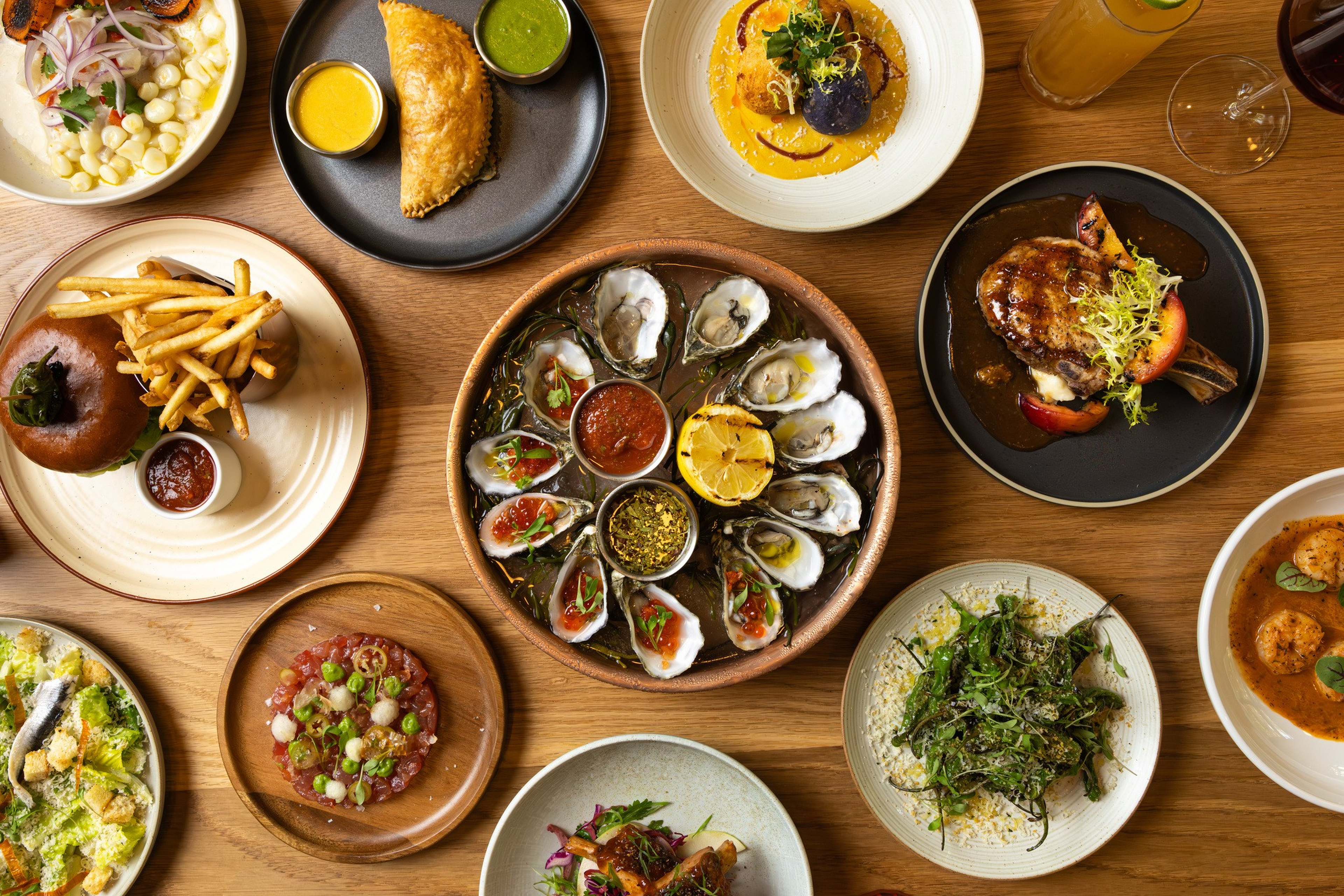An array of dishes includes oysters with sauces, a burger with fries, empanada with green and yellow sauces, grilled meat with greens, seafood in sauce, and fresh salads.