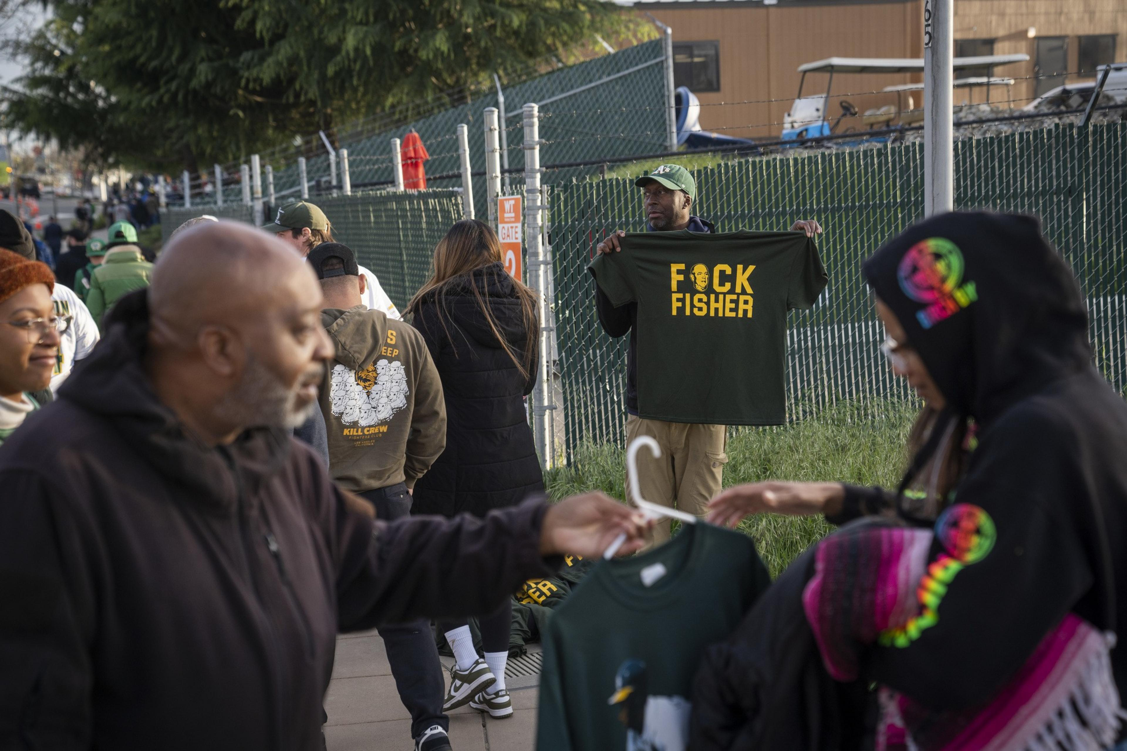 People stand near a fence at an outdoor market, including a man holding a green shirt with yellow lettering. Others browse nearby merchandise, with trees in the background.