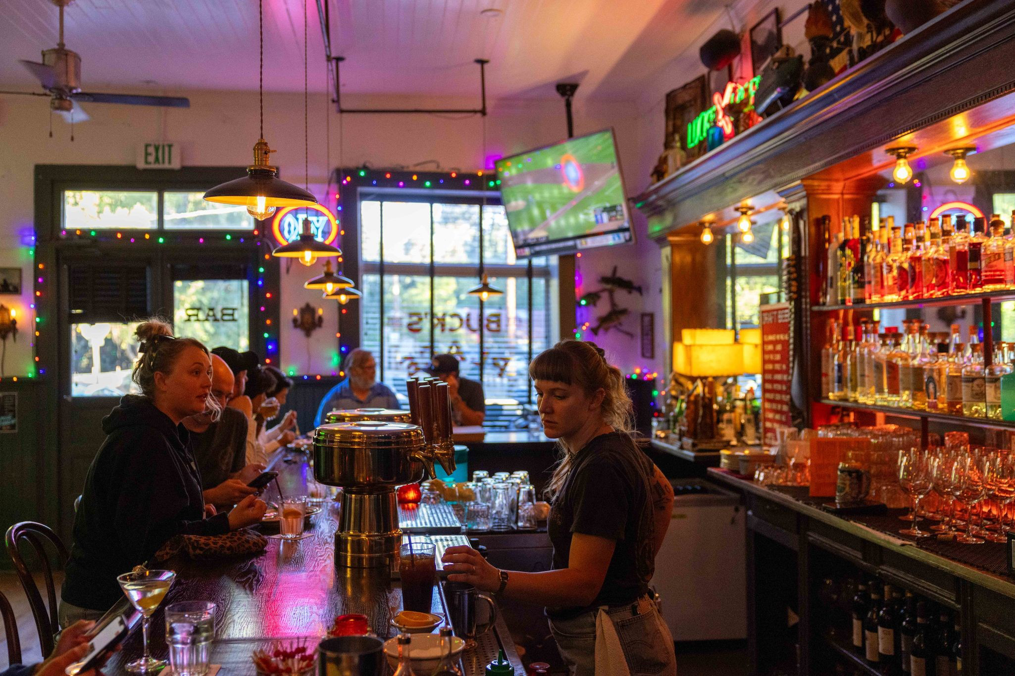 A bartender serves drinks at a cozy bar with customers sitting along the wooden counter and colorful string lights decorating the space.