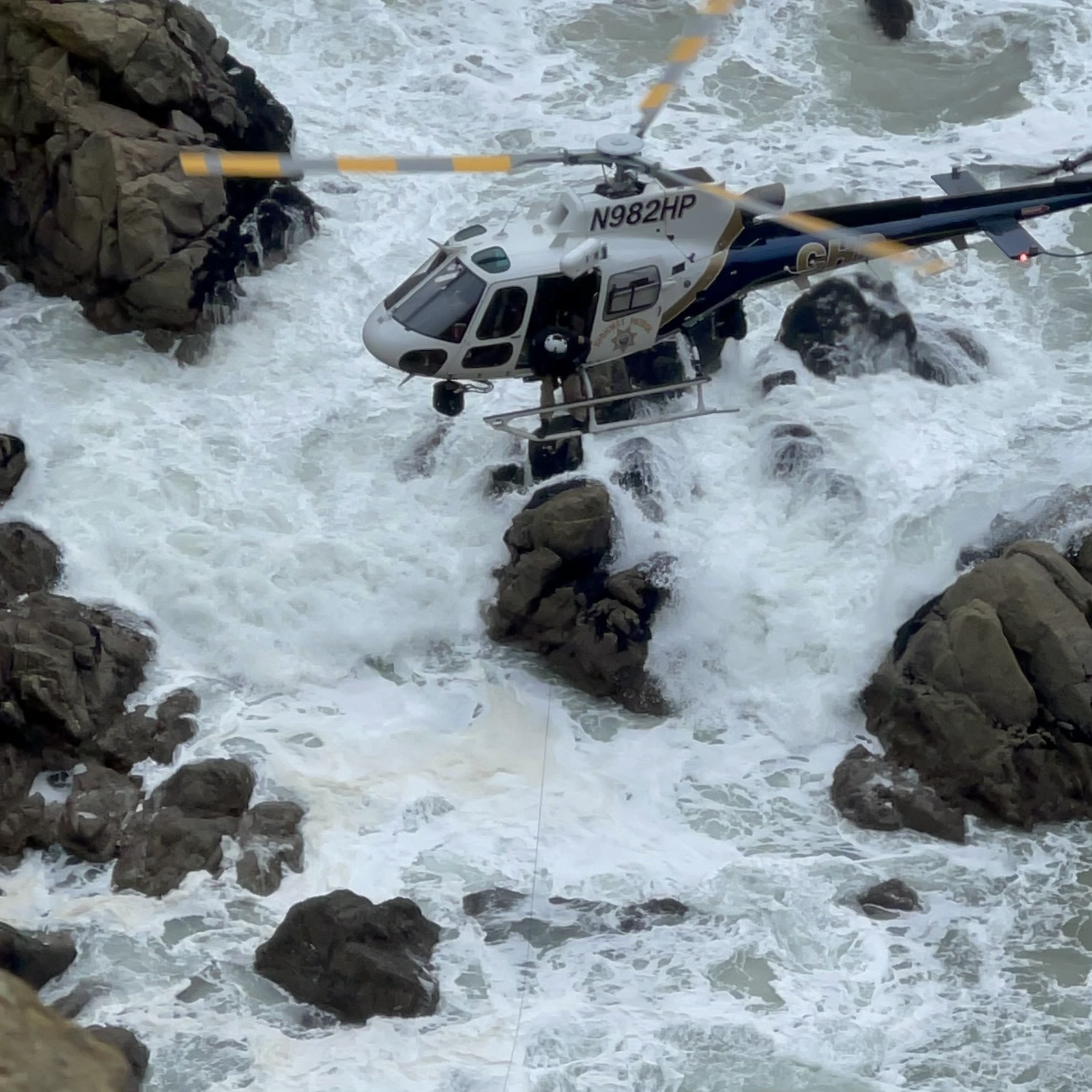 A helicopter hovers above turbulent ocean waves and rocks, with a person visible near the open side door and a cable extending downward.
