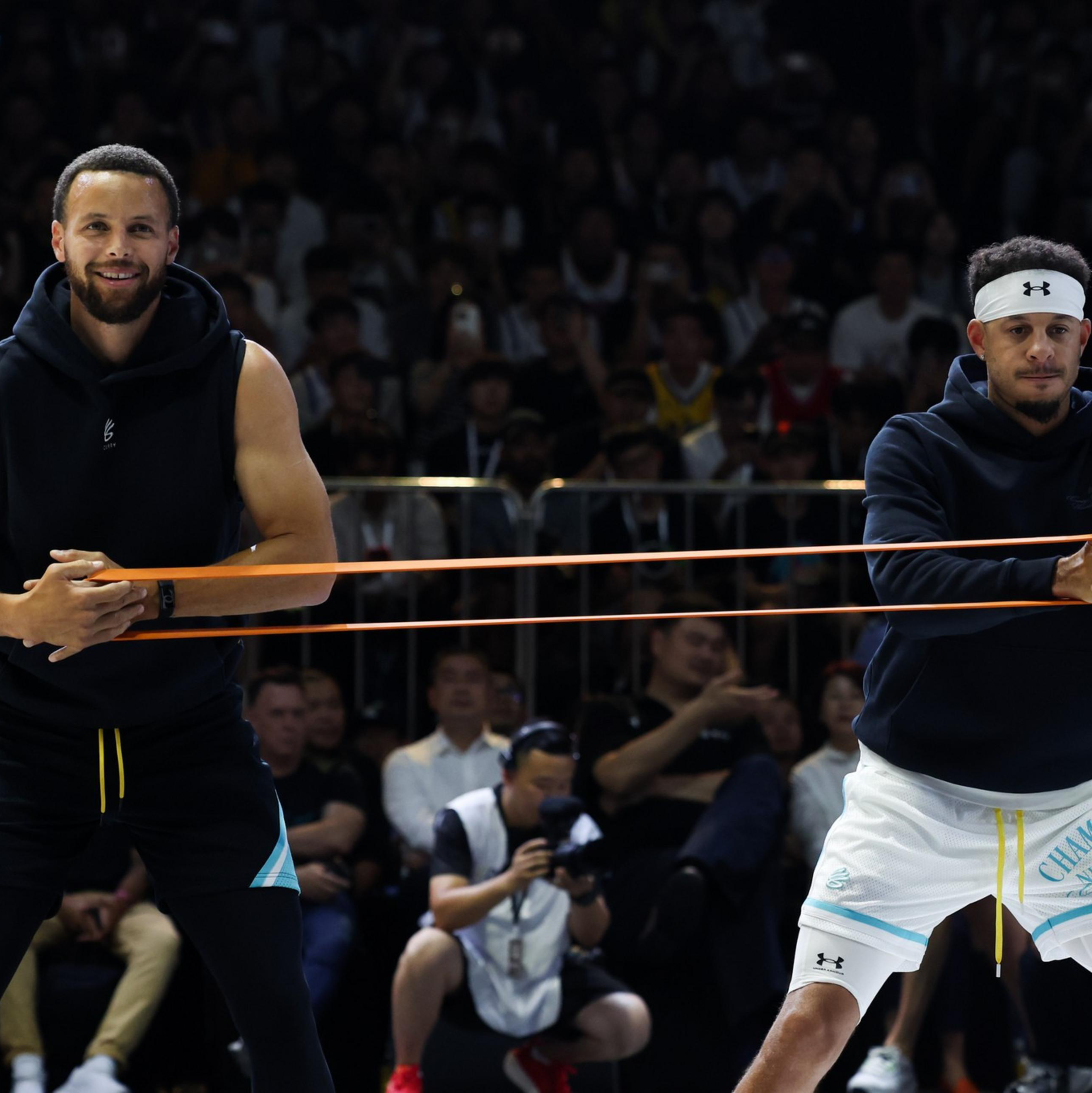 Two athletes stretch an orange resistance band between them while standing in a sports arena filled with spectators.