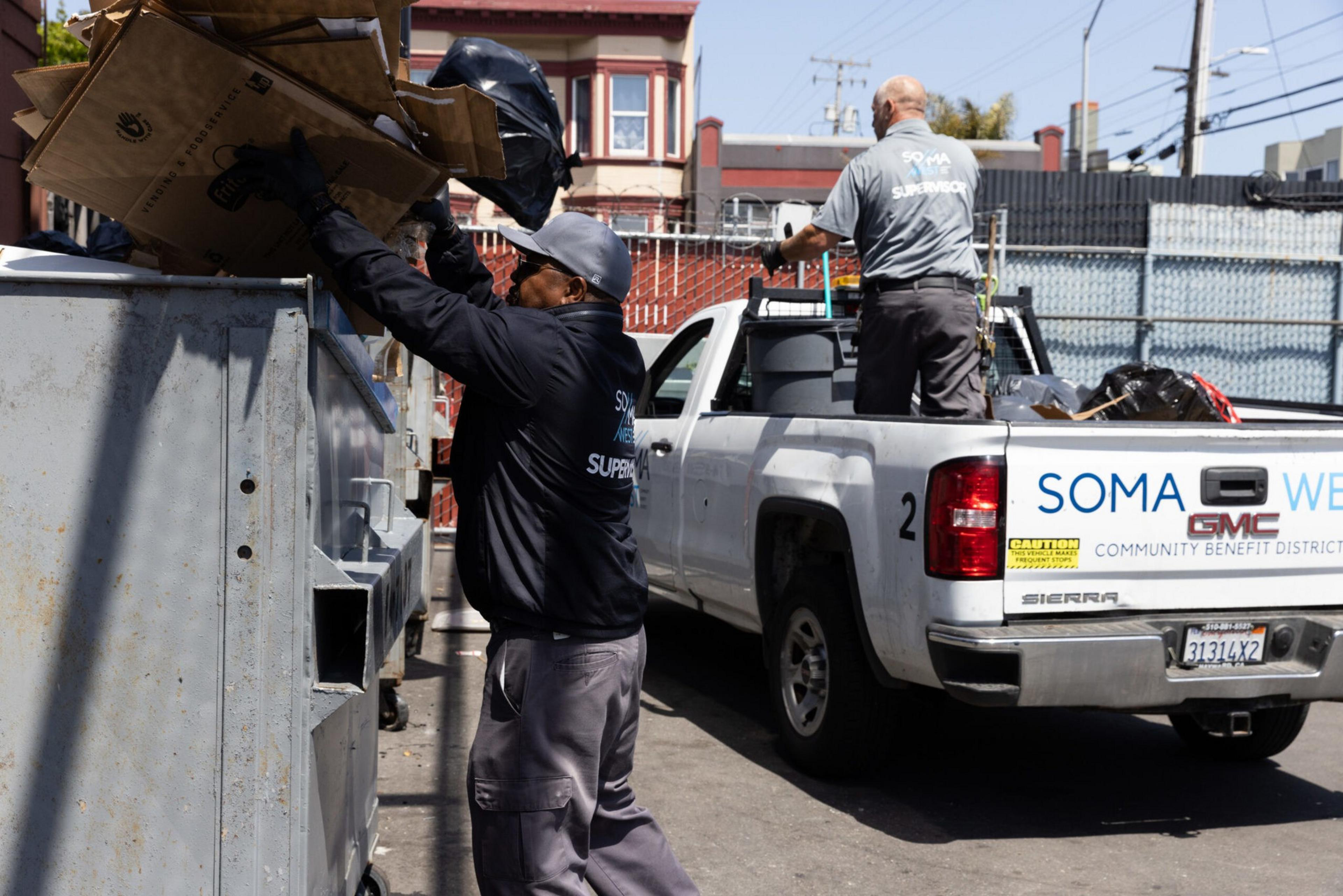 A man wearing a cap and jacket loads cardboard and bags into a large gray dumpster while another man stands on the back of a white pickup truck nearby.