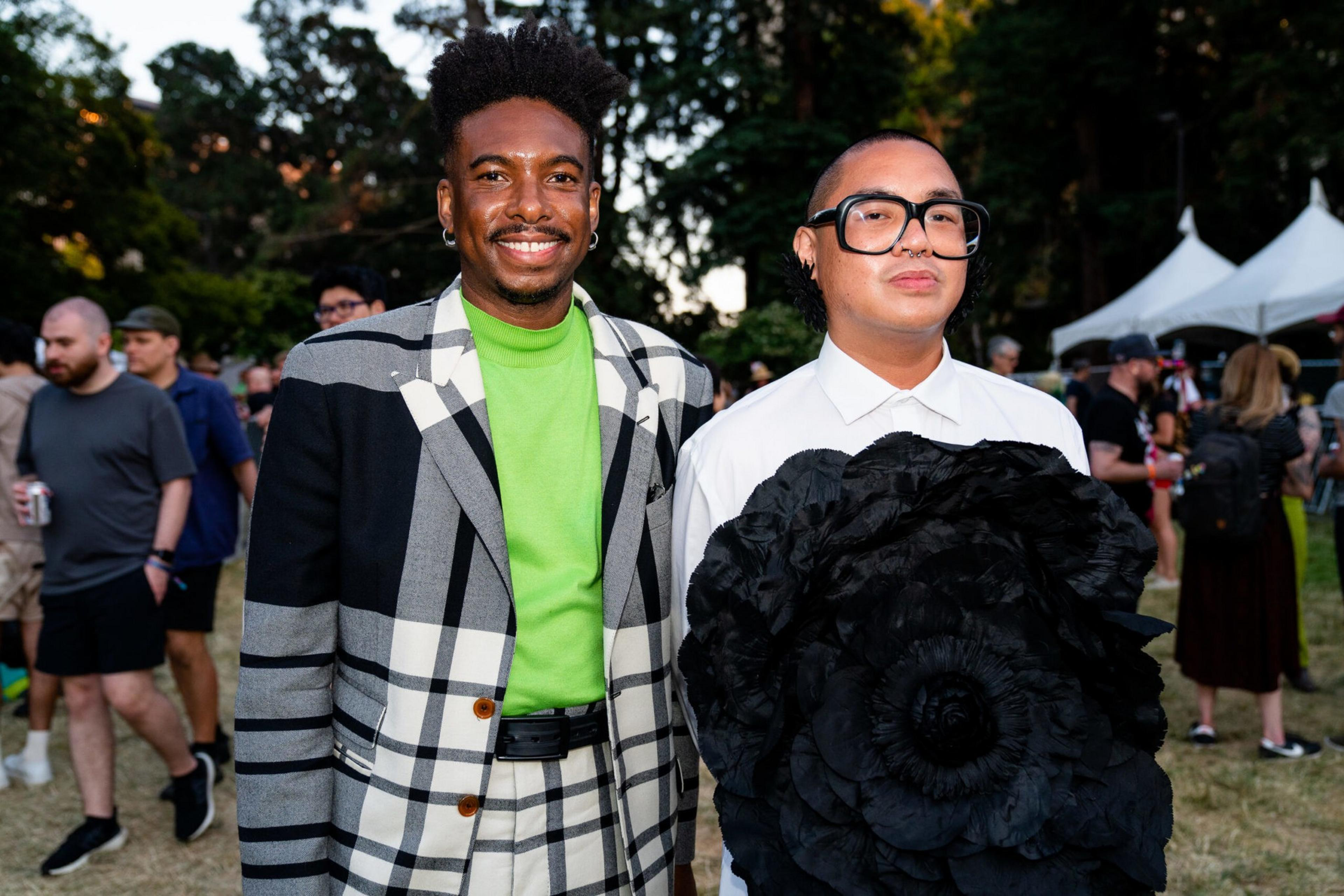 Two men stand outdoors at an event; one wears a green shirt with a black-and-white plaid suit, the other wears a white shirt and large black glasses holding a large black flower.