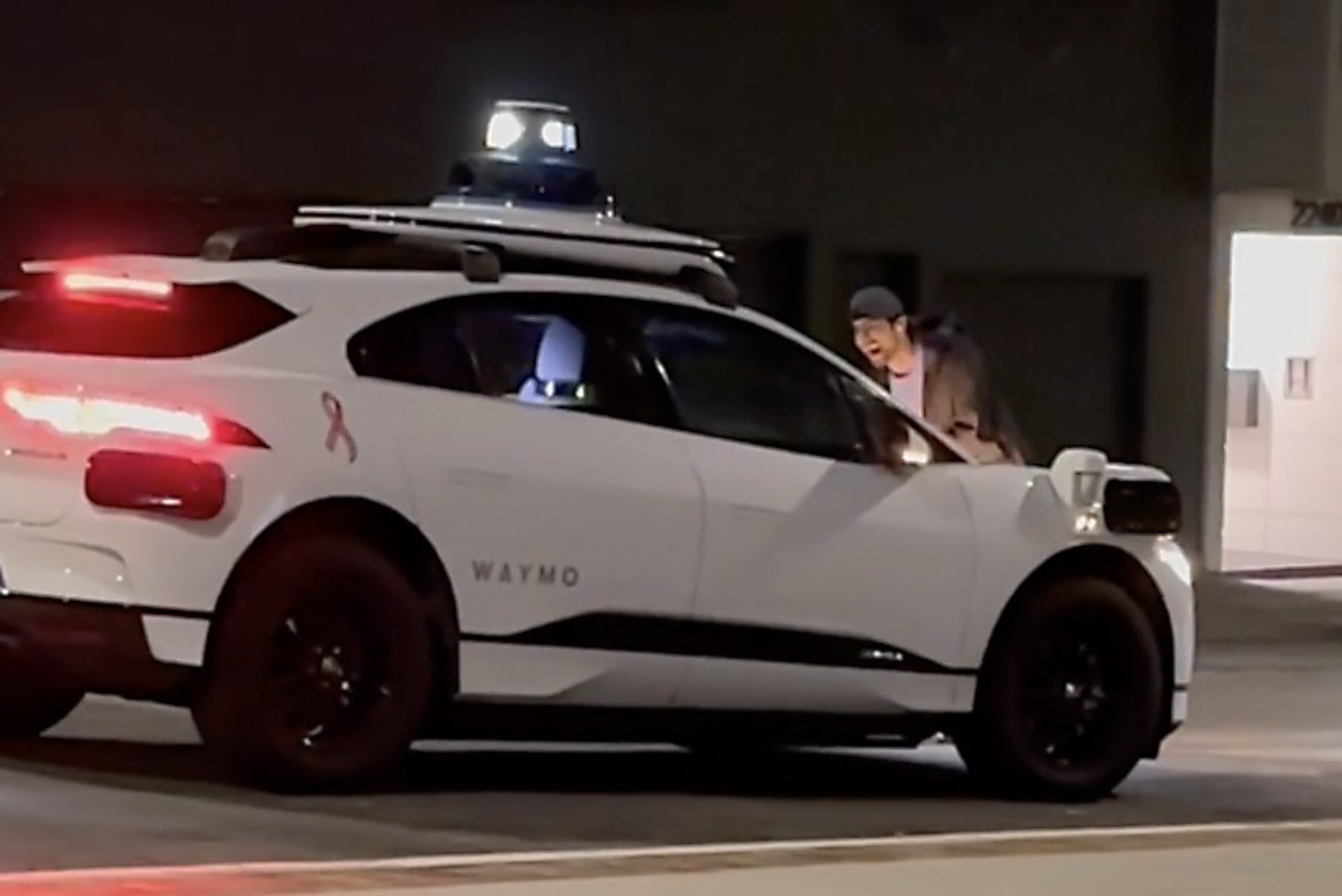 A white Waymo autonomous vehicle with a roof sensor is parked on the street at night, and a person is standing next to the driver's window, looking inside.