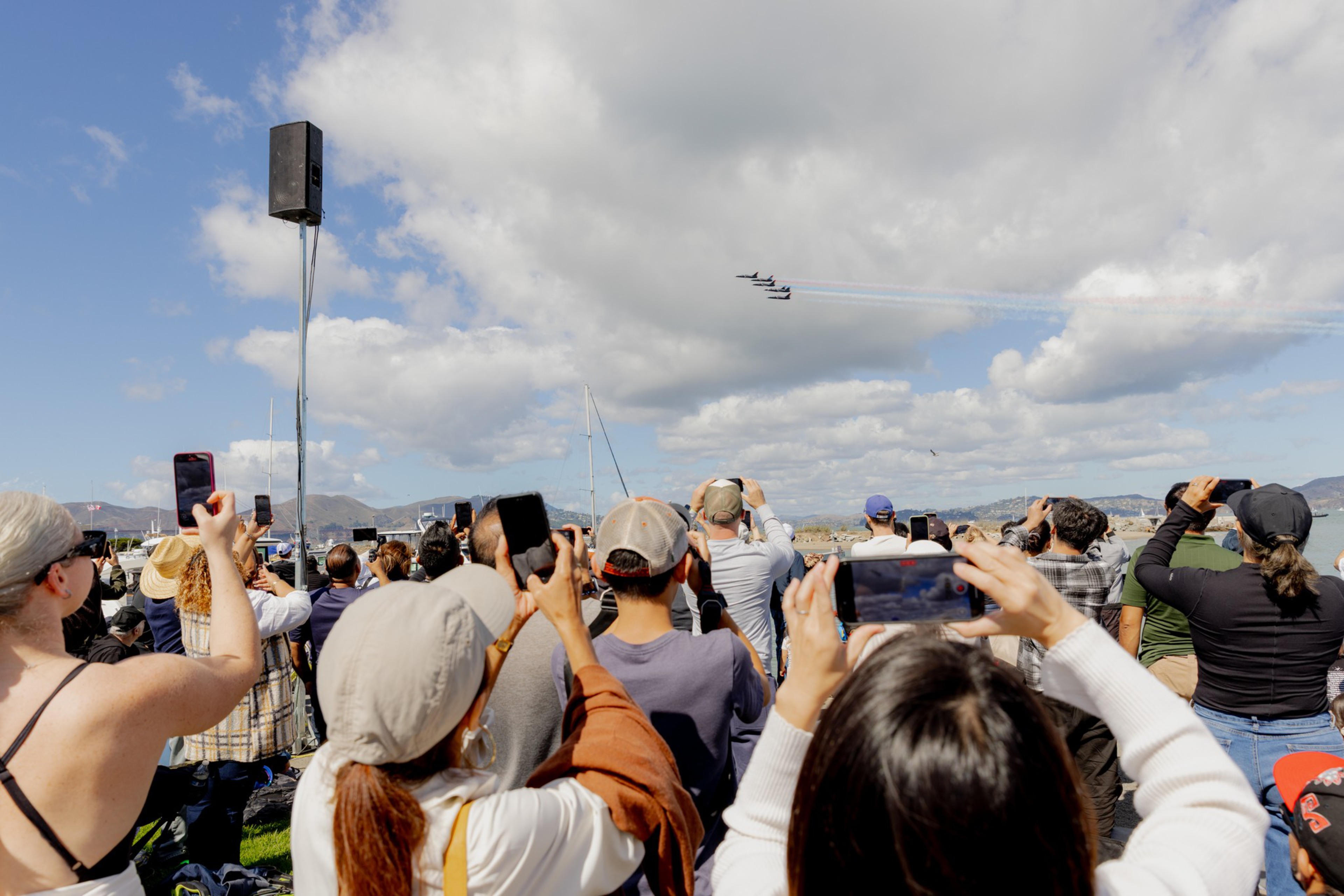 A crowd of people outdoors is watching and recording an airshow with four jets flying in tight formation against a partly cloudy sky.