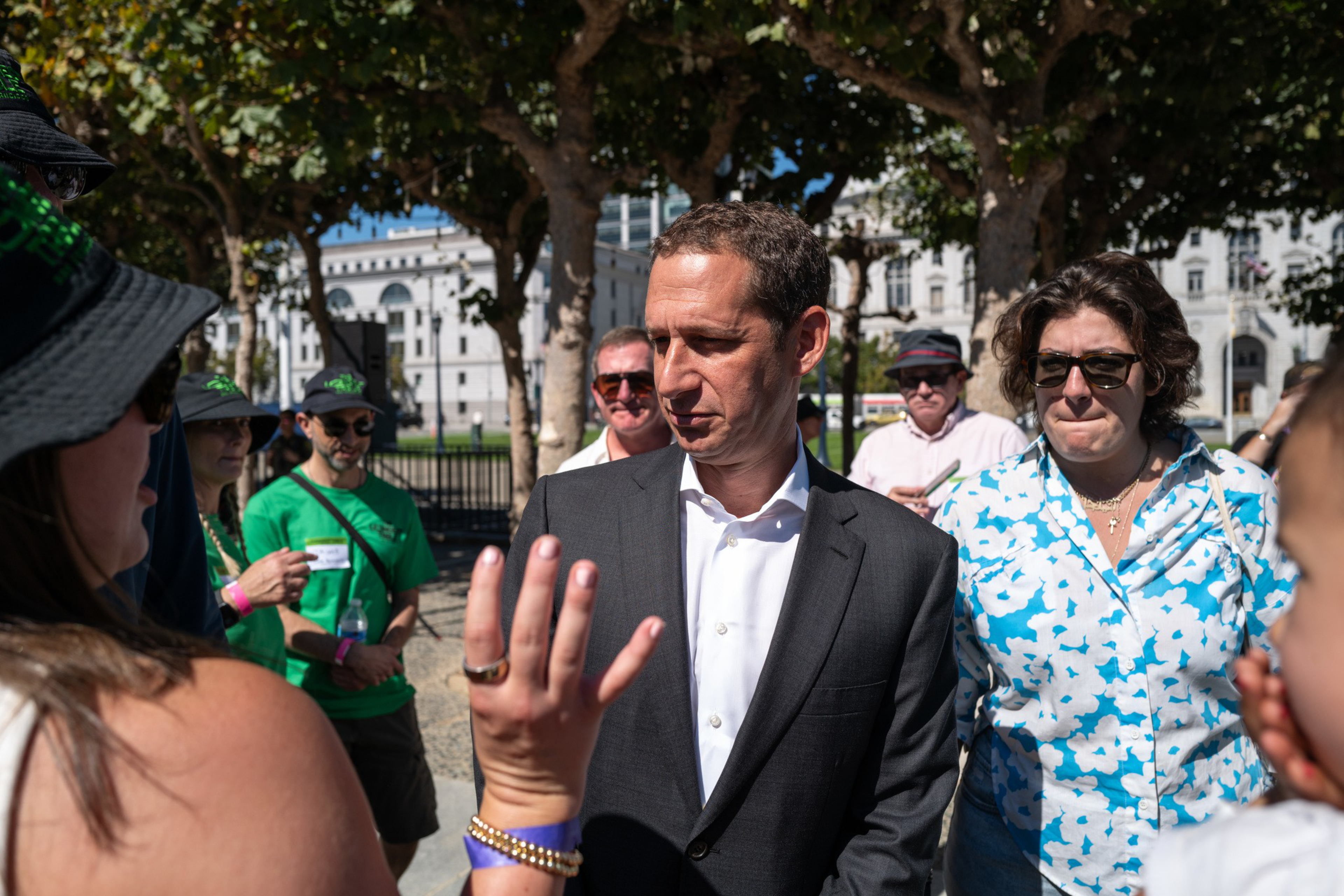 A man in a dark suit listens to a woman gesturing with her hand, surrounded by casually dressed people outdoors near trees and buildings.