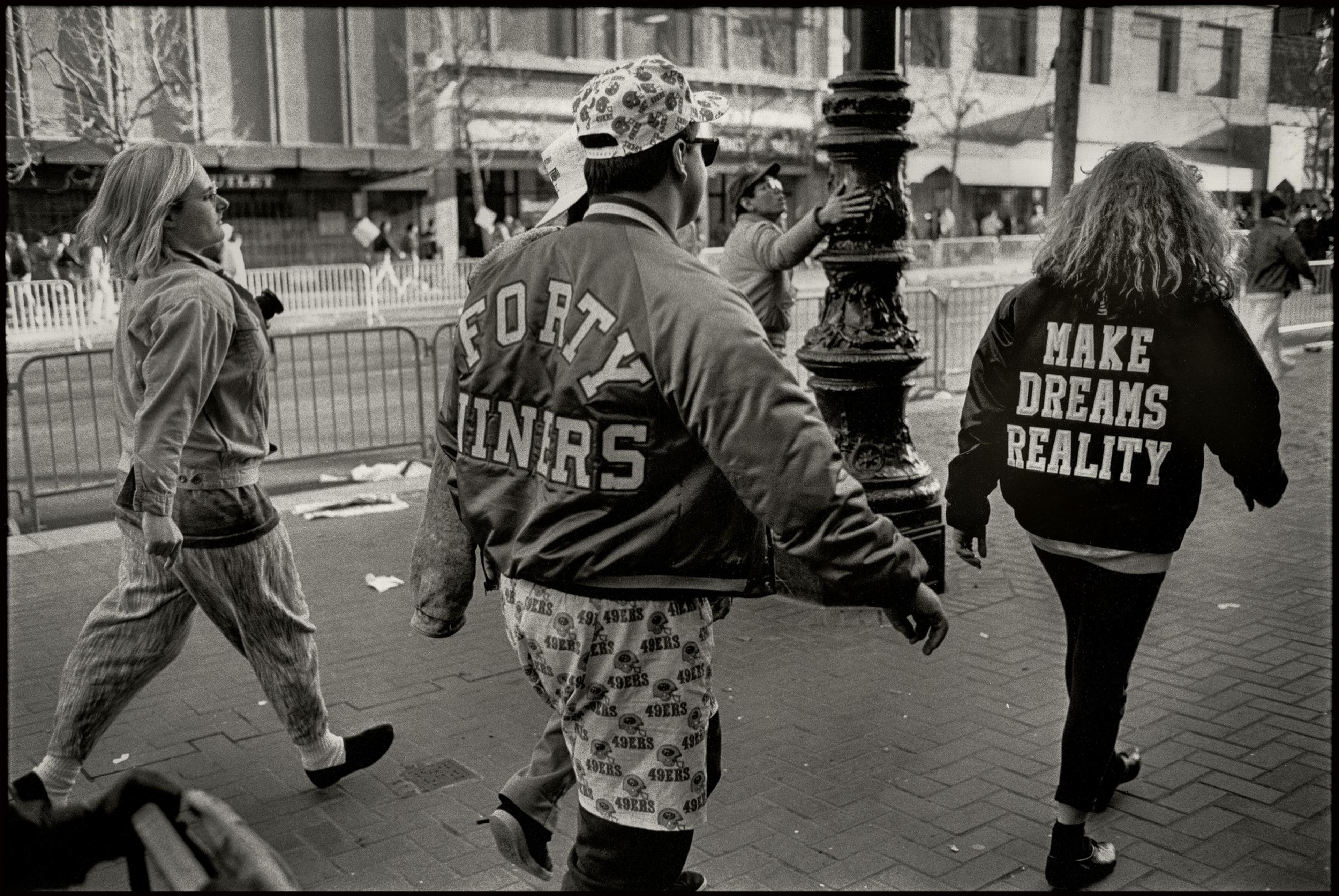 A black and white photo of a group of people, one wearing a vintage Forty-Niners jacket, walk along a street.