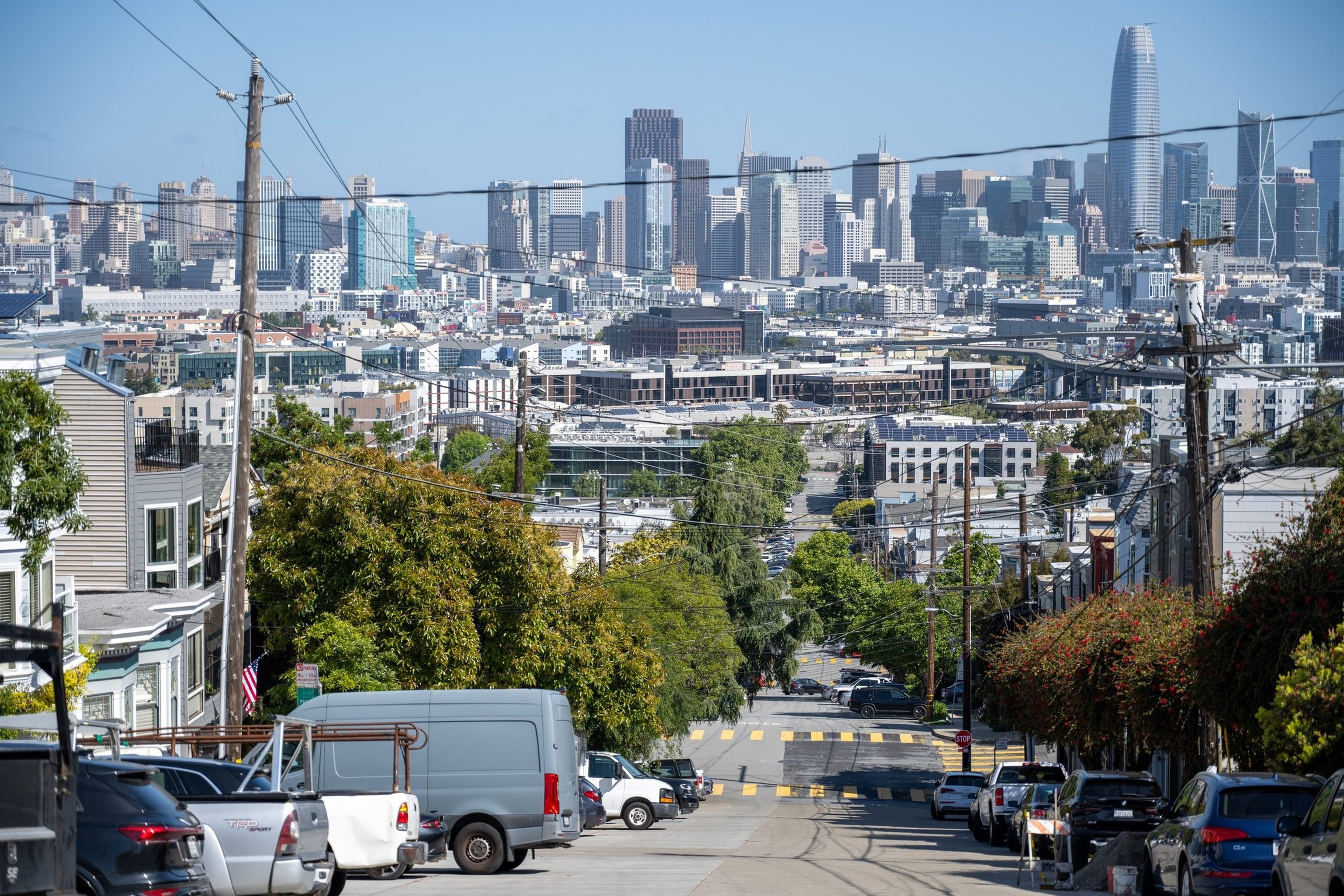 A sloped city street lined with parked cars and trees leads toward a distant urban skyline filled with tall buildings under a clear blue sky.