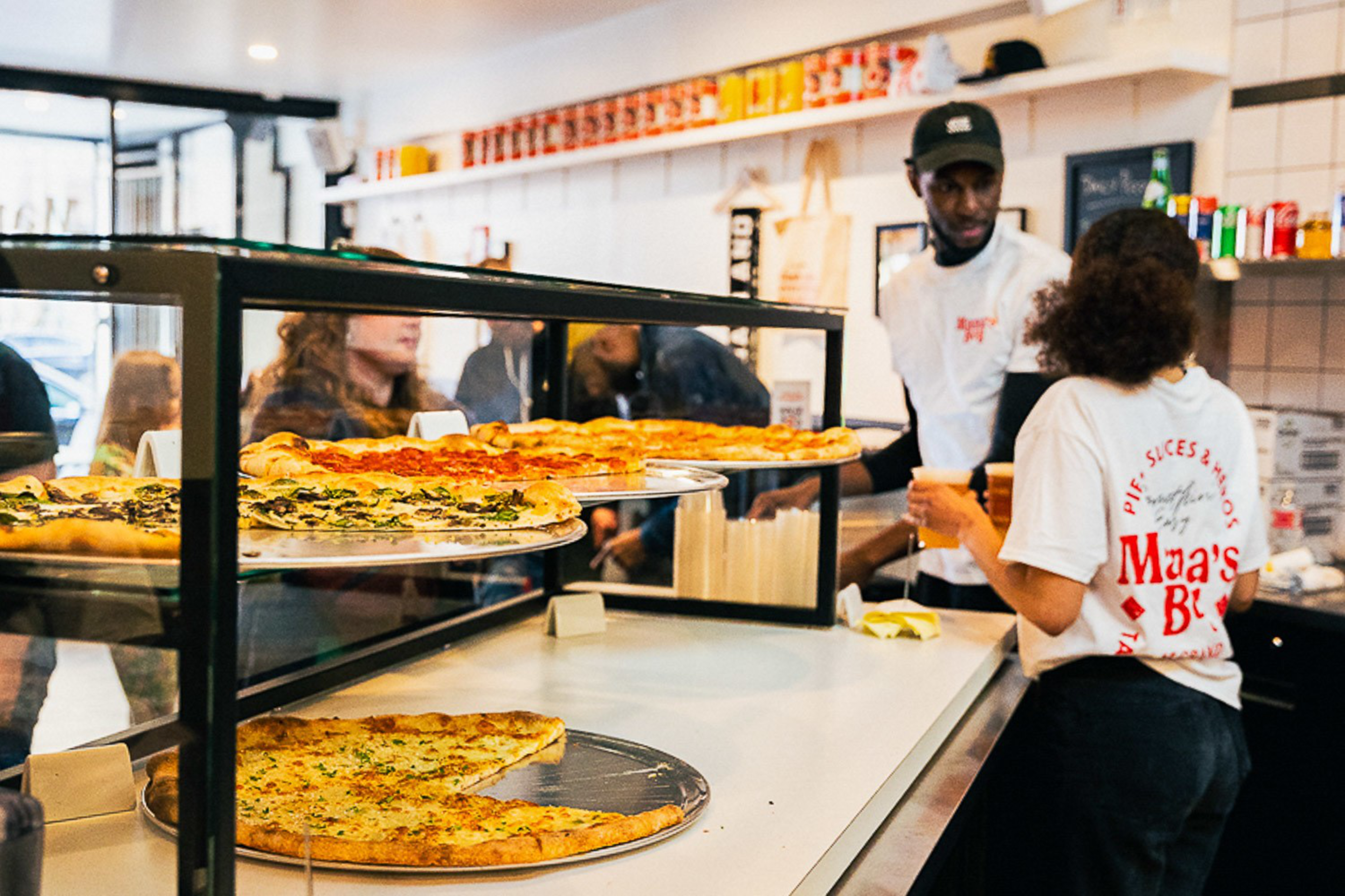 A pizza counter displays multiple large slices while two employees in white shirts serve customers in a busy, brightly lit pizzeria.