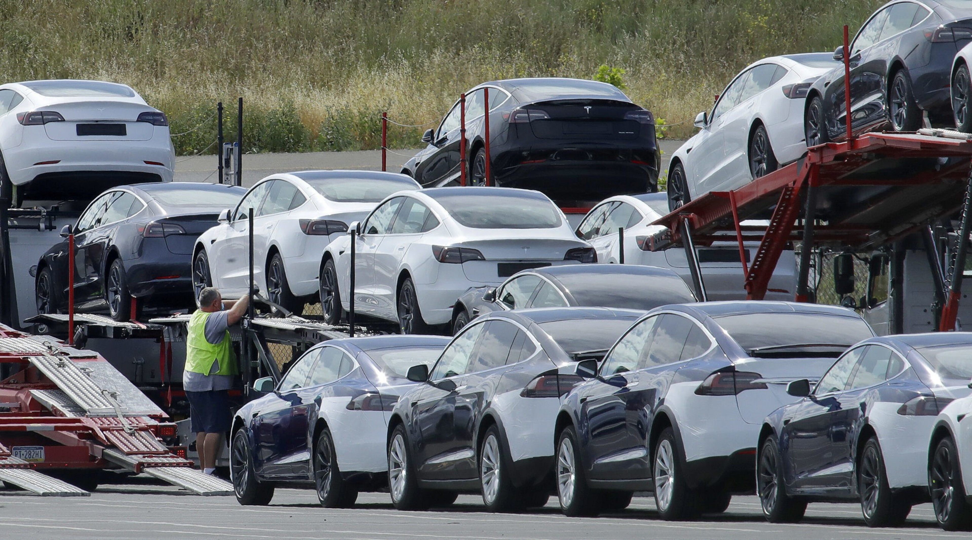 A person in a reflective vest is loading or unloading new cars from a transporter truck in a parking lot.