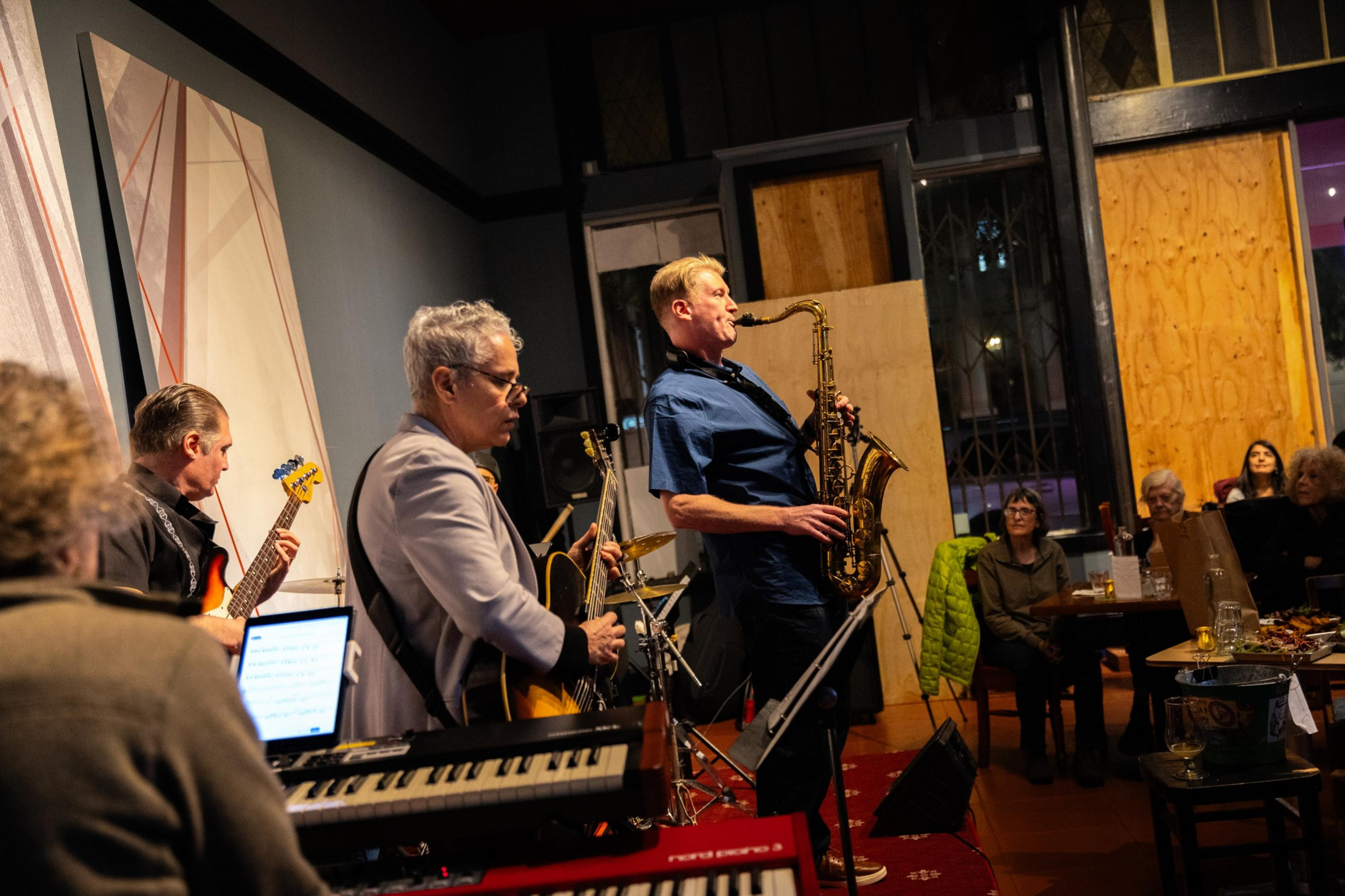 A band performing indoors with a saxophonist in focus; audience watching in the background.