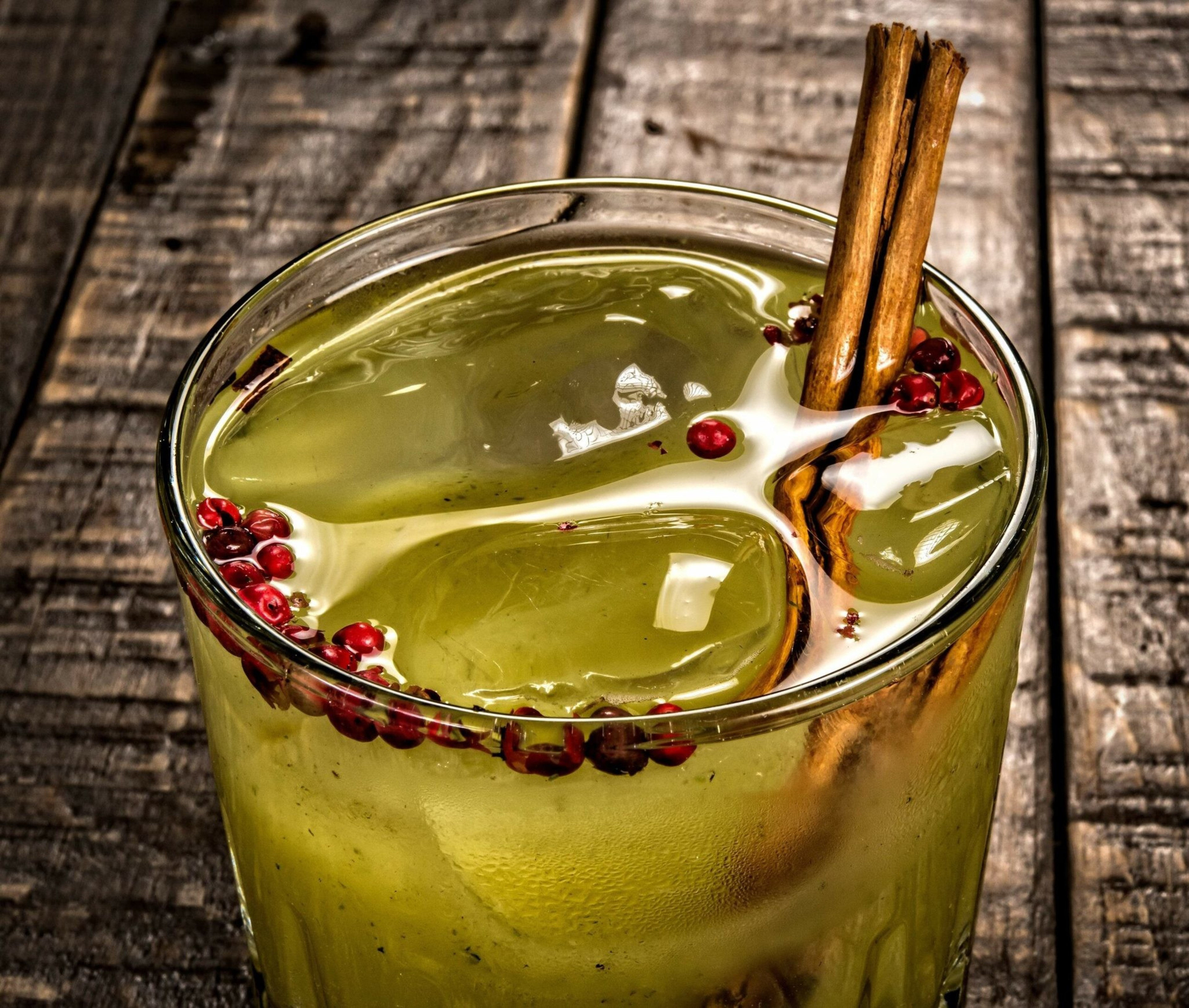 A glass of yellow-green drink with ice cubes, two cinnamon sticks, and small red peppercorns floating on top, placed on a wooden surface.