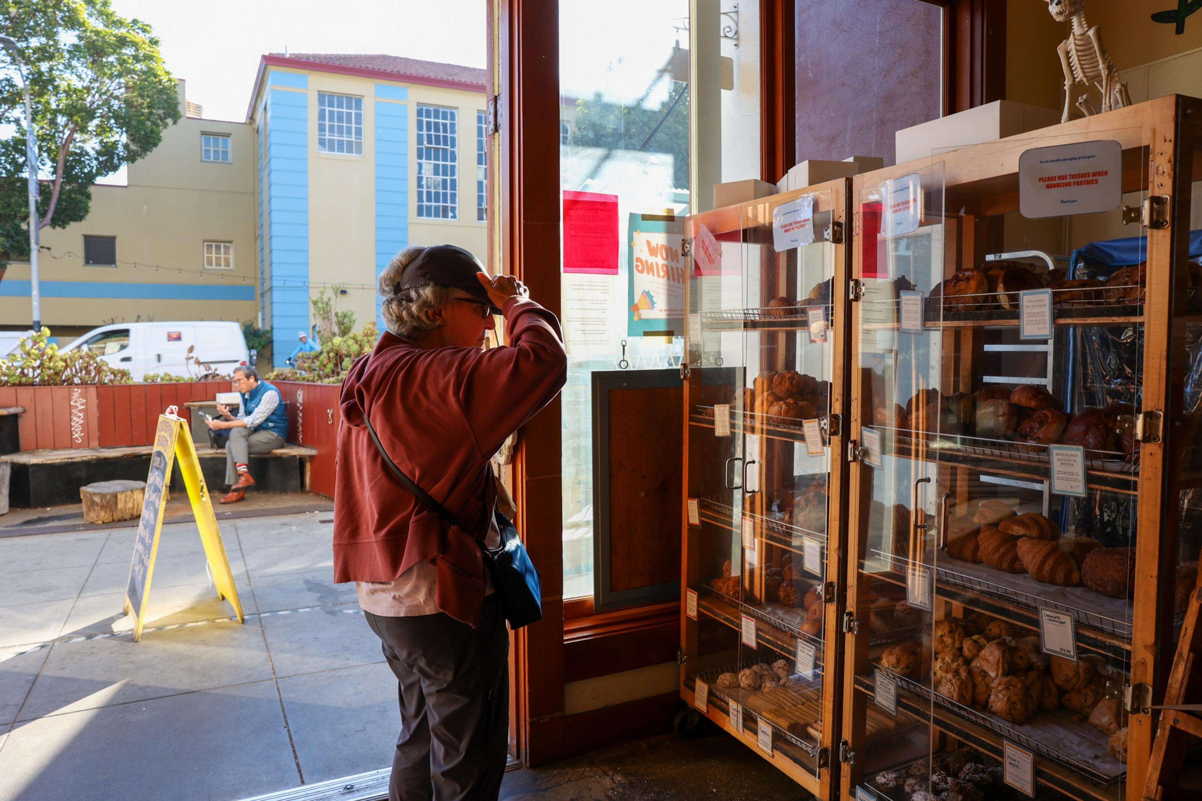 A person wearing a cap and red jacket looks at bakery goods inside a glass display near a sunlit entrance with another person sitting outside.