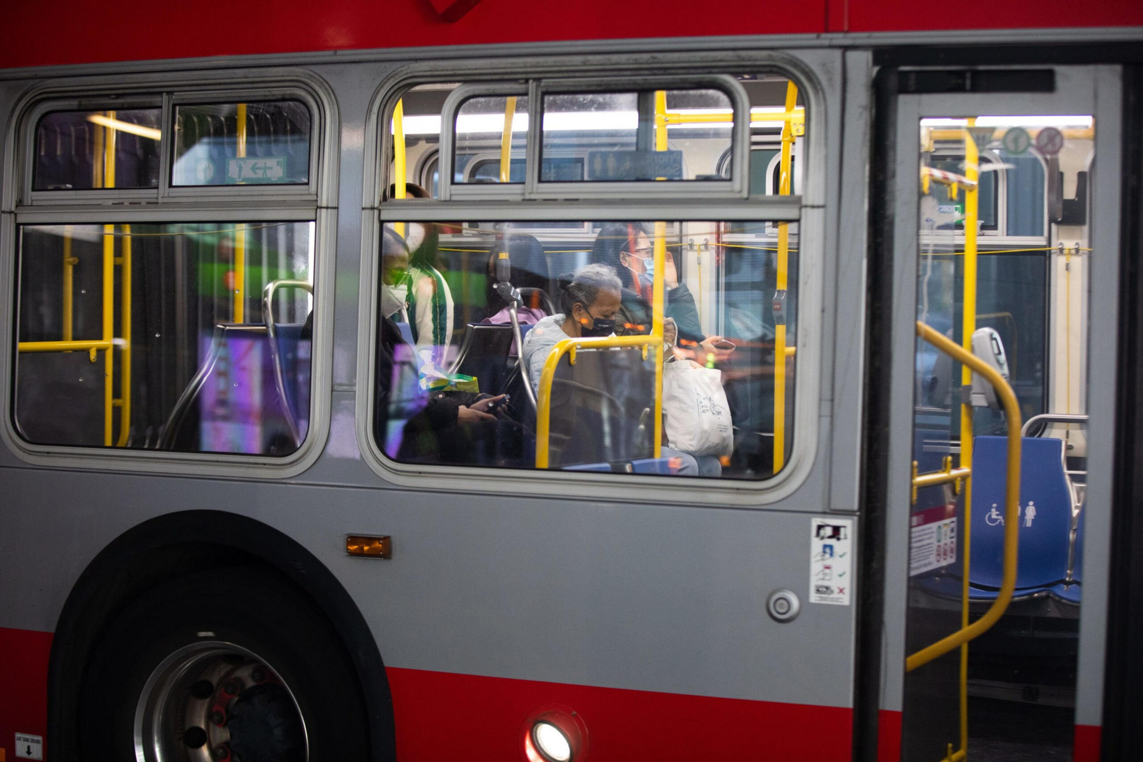 Several passengers wearing masks sit inside a red and gray bus, some looking at their phones, with yellow handrails and a wheelchair-accessible seat visible.