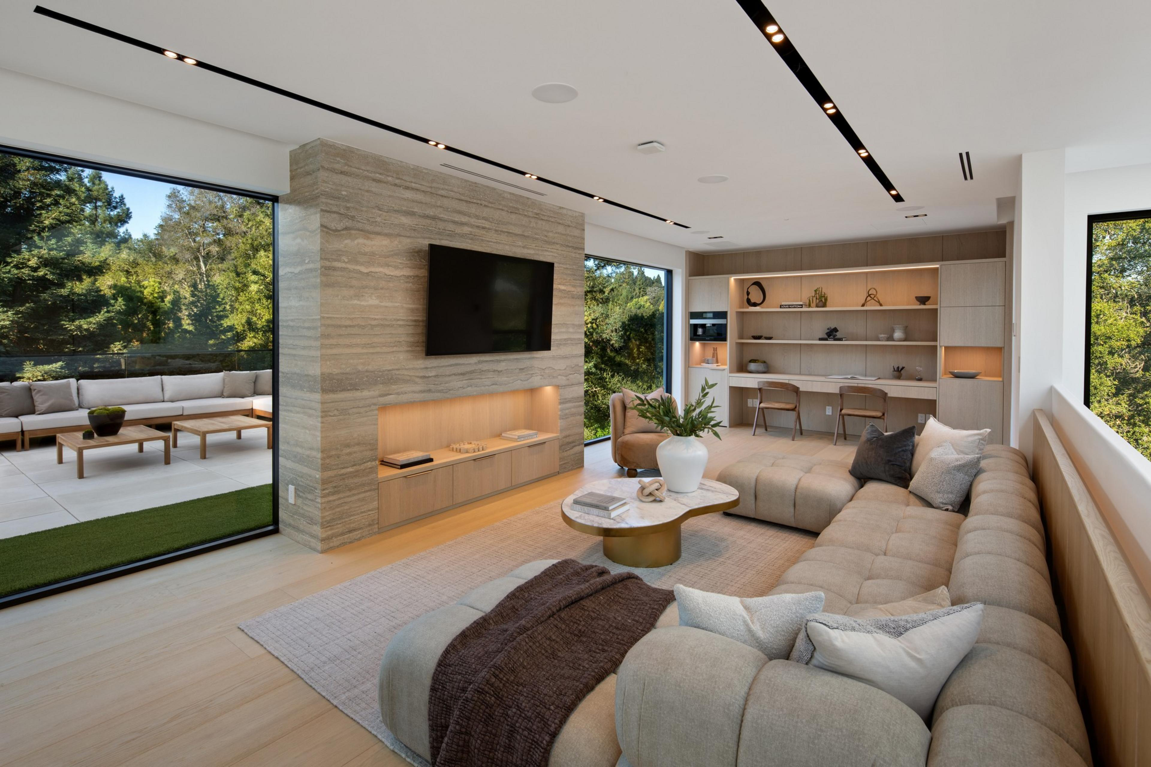 A modern living room features a large beige sectional sofa, a round marble coffee table, built-in shelves with decor, and a wall-mounted TV, with large windows overlooking greenery.