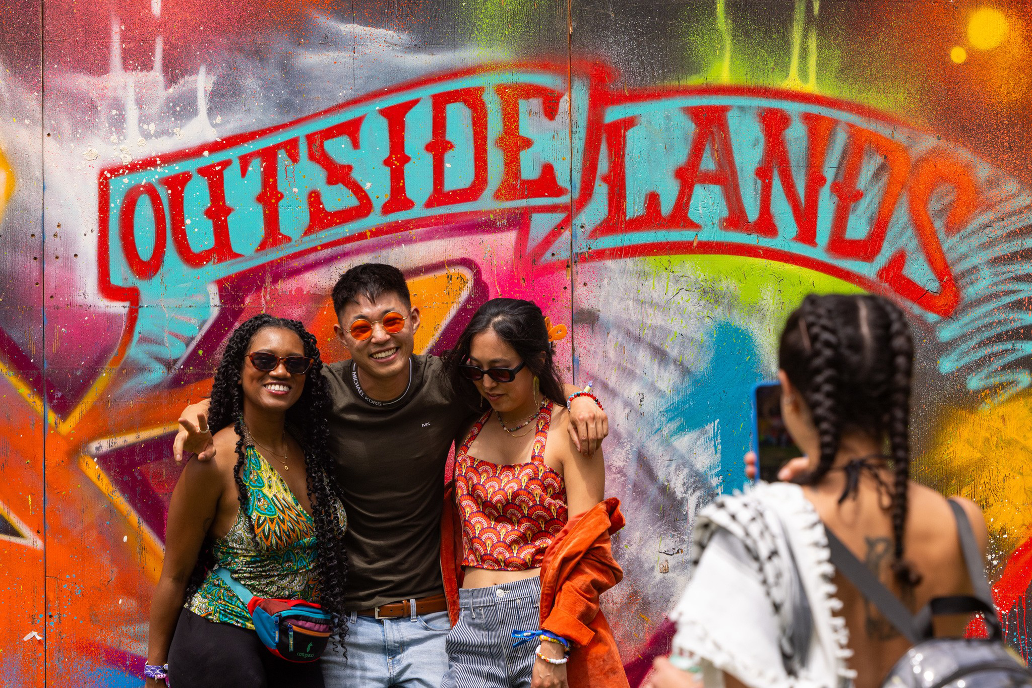 Three friends pose smiling in front of a colorful graffiti wall that says "OUTSIDE LANDS" while a fourth person takes their photo.