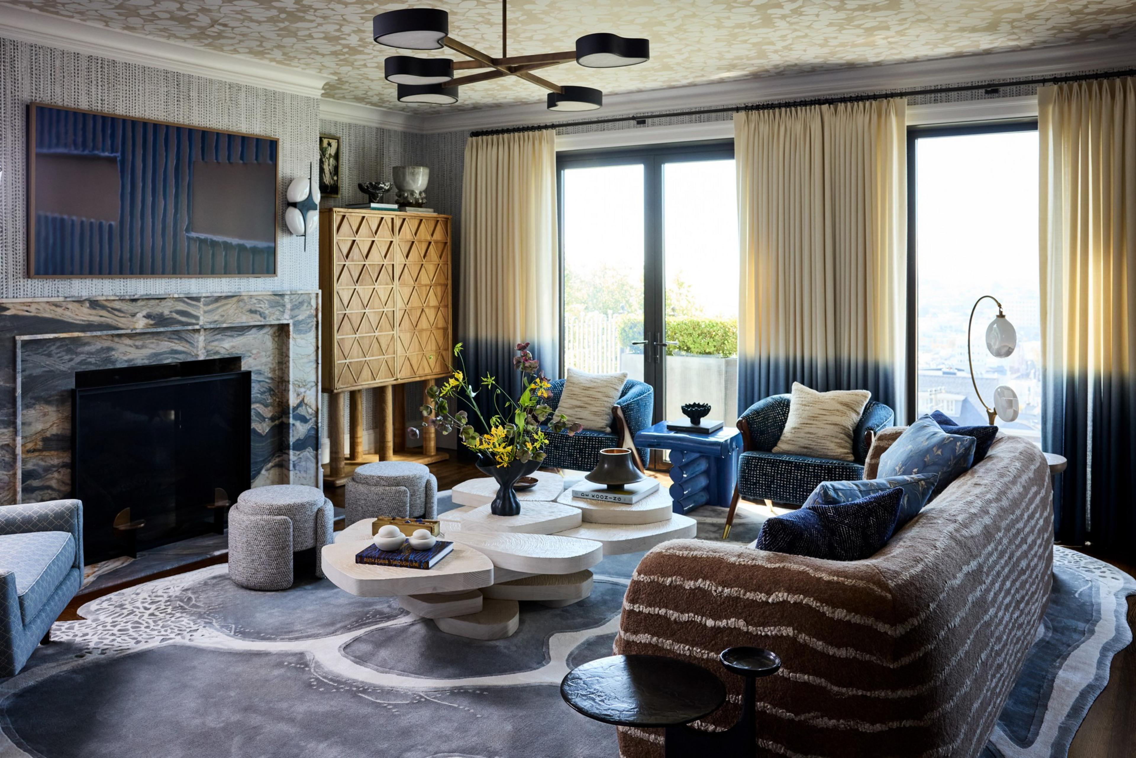 A cozy living room with patterned beige and brown sofa, blue armchairs, layered white coffee tables, textured rug, large windows with beige and blue curtains, and a marble fireplace.