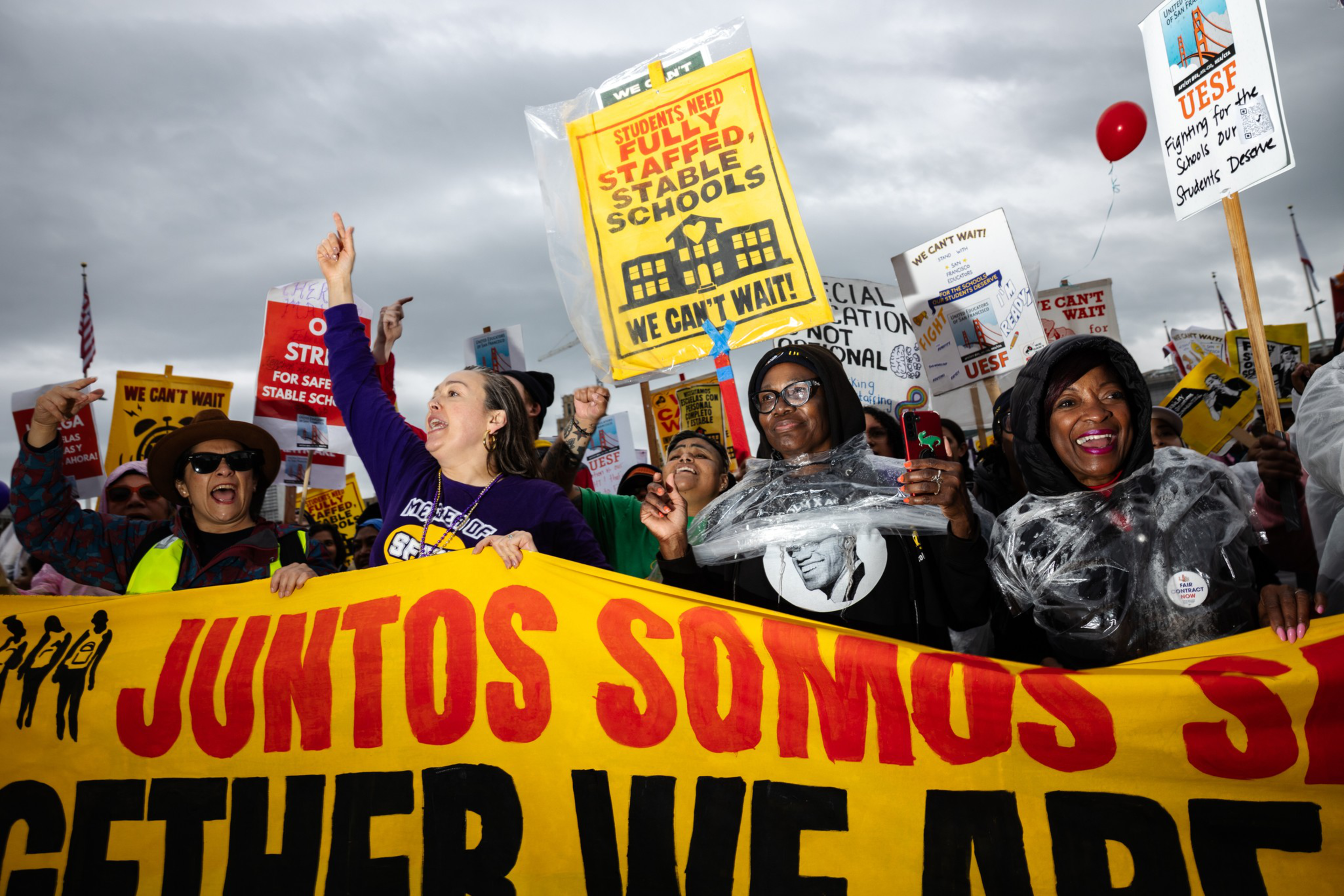 A diverse group of people hold a large yellow banner with red and black text at a protest, carrying signs demanding fully staffed, stable schools.