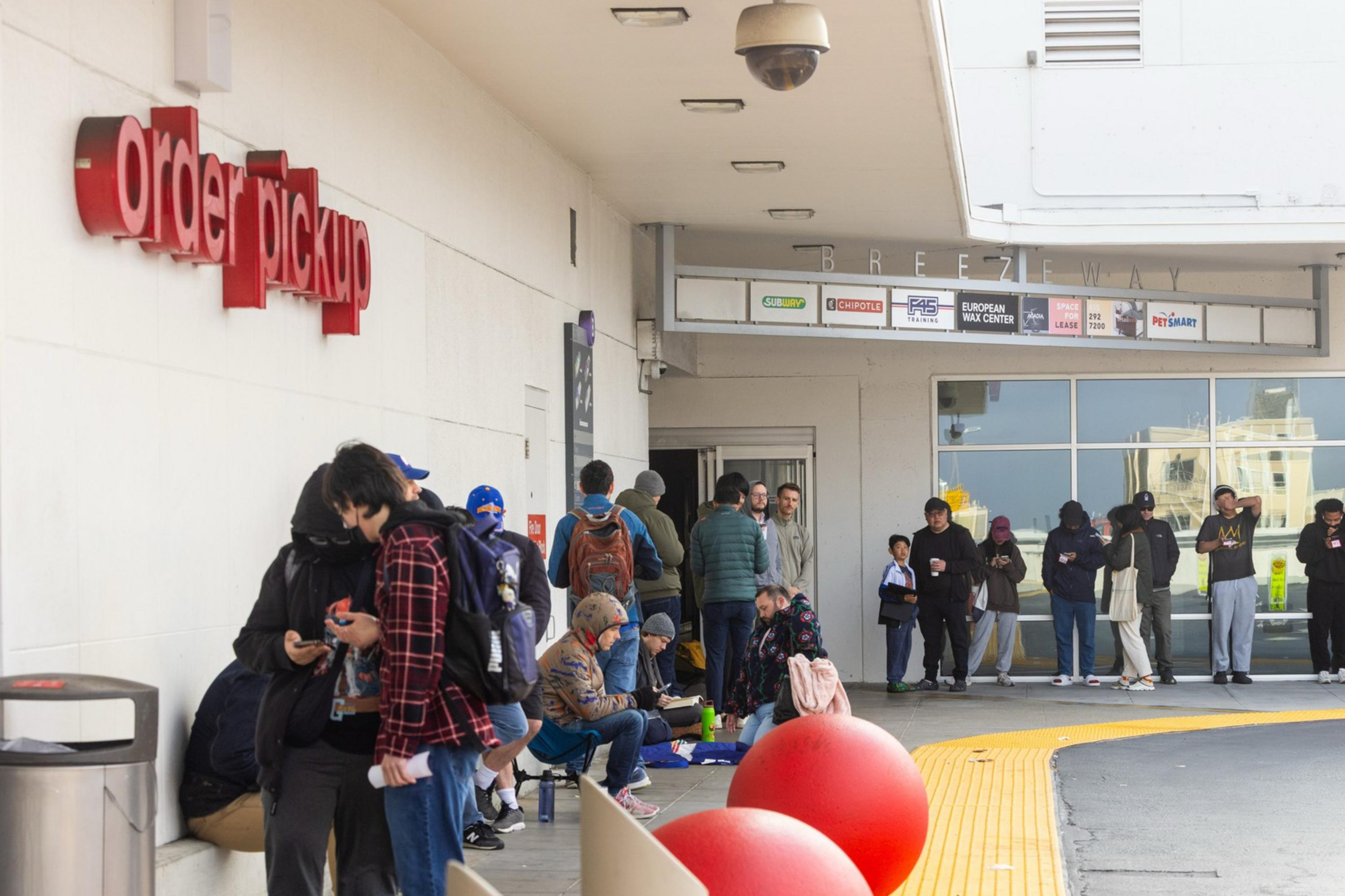 A group of people are lined up outside an "order pickup" area, some standing and others sitting, surrounded by large red spheres and a trash can.