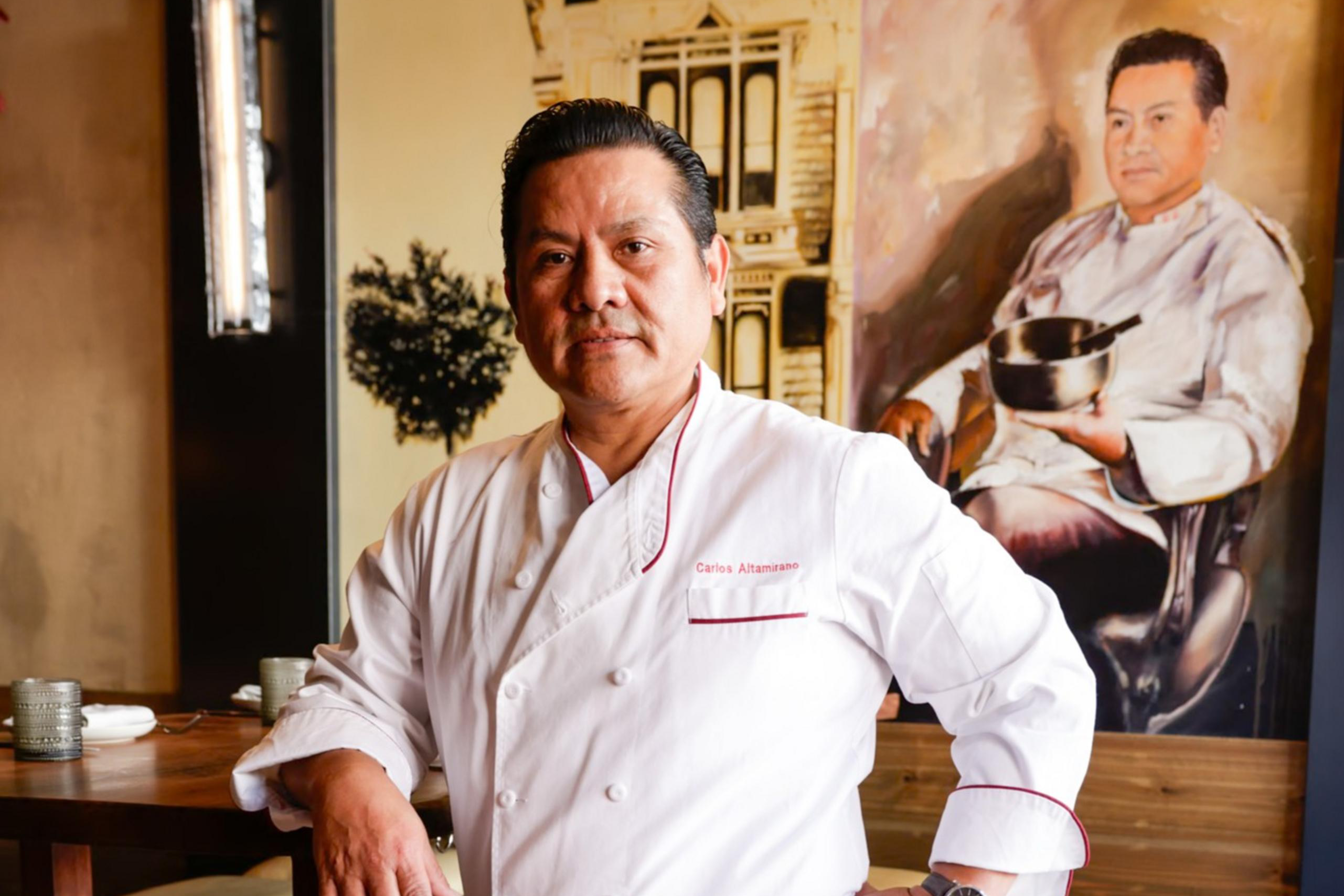 A chef in a white coat stands confidently beside a wooden table. Behind him, there's a portrait featuring the chef holding a pot, with a vintage building in the background.