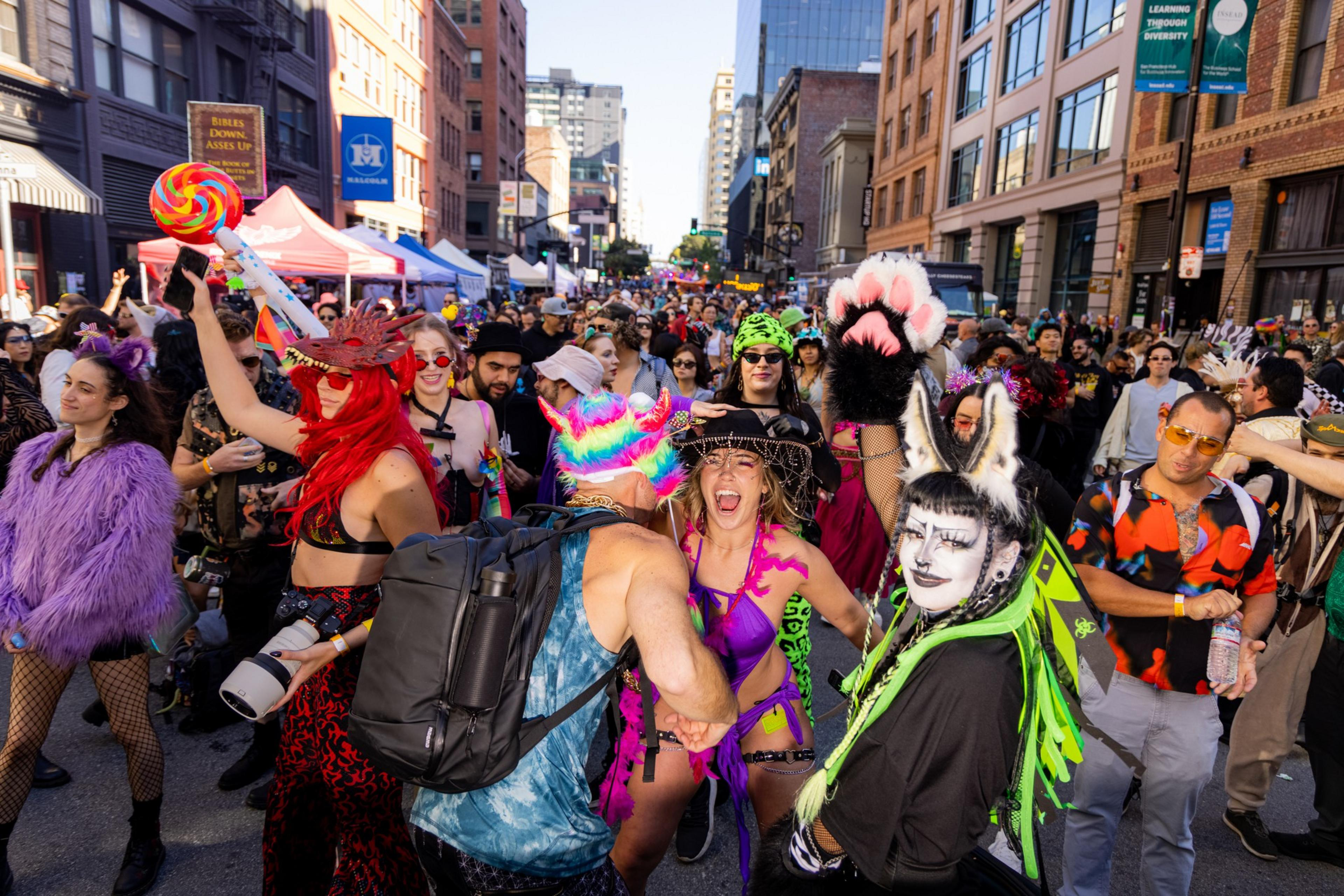 A lively street filled with people in colorful, eclectic costumes and accessories, celebrating in high spirits. Buildings and tents line the background.