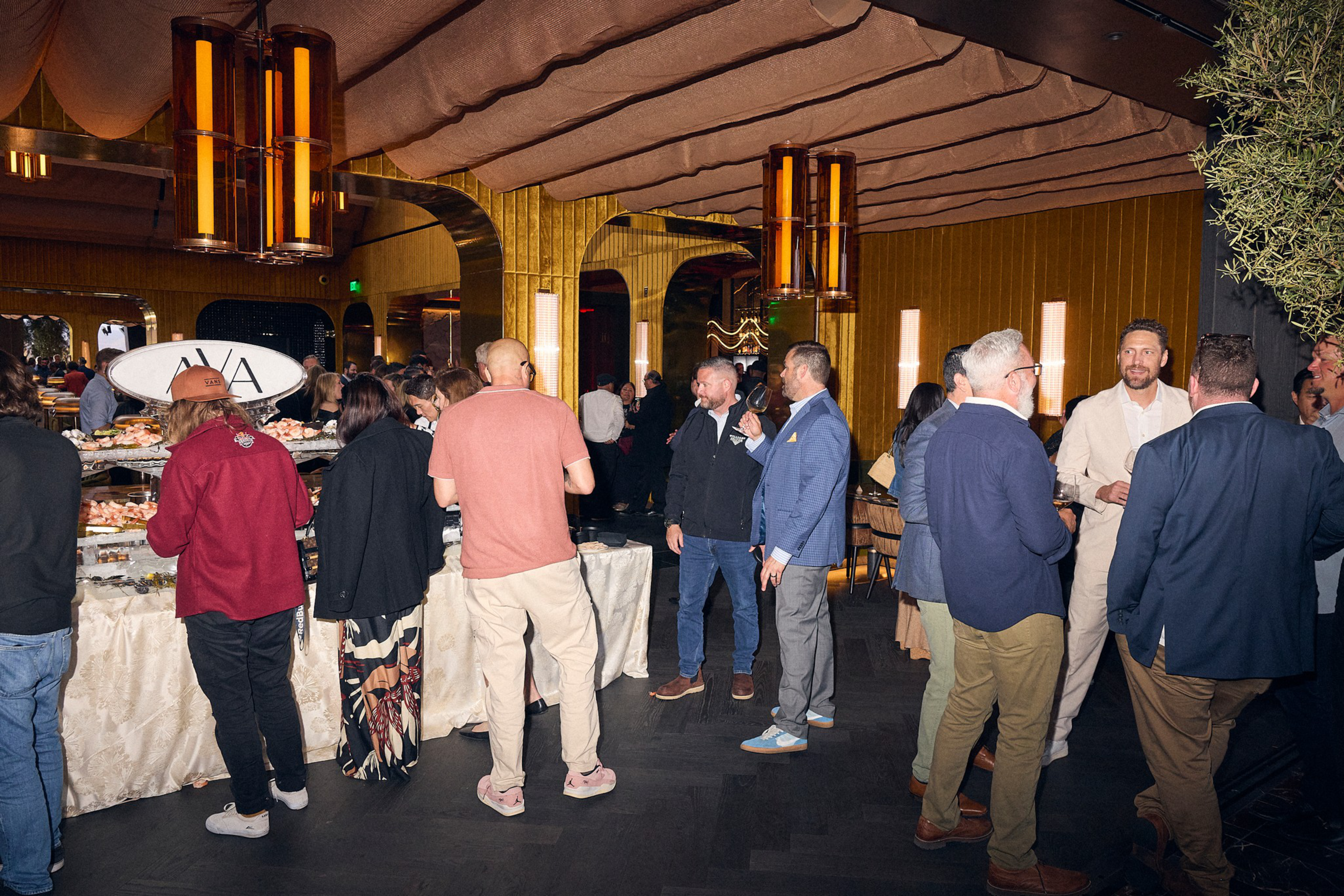 A group of people are dressed in casual and semi-formal attire, socializing and standing around a buffet table with food in a warmly lit indoor venue.