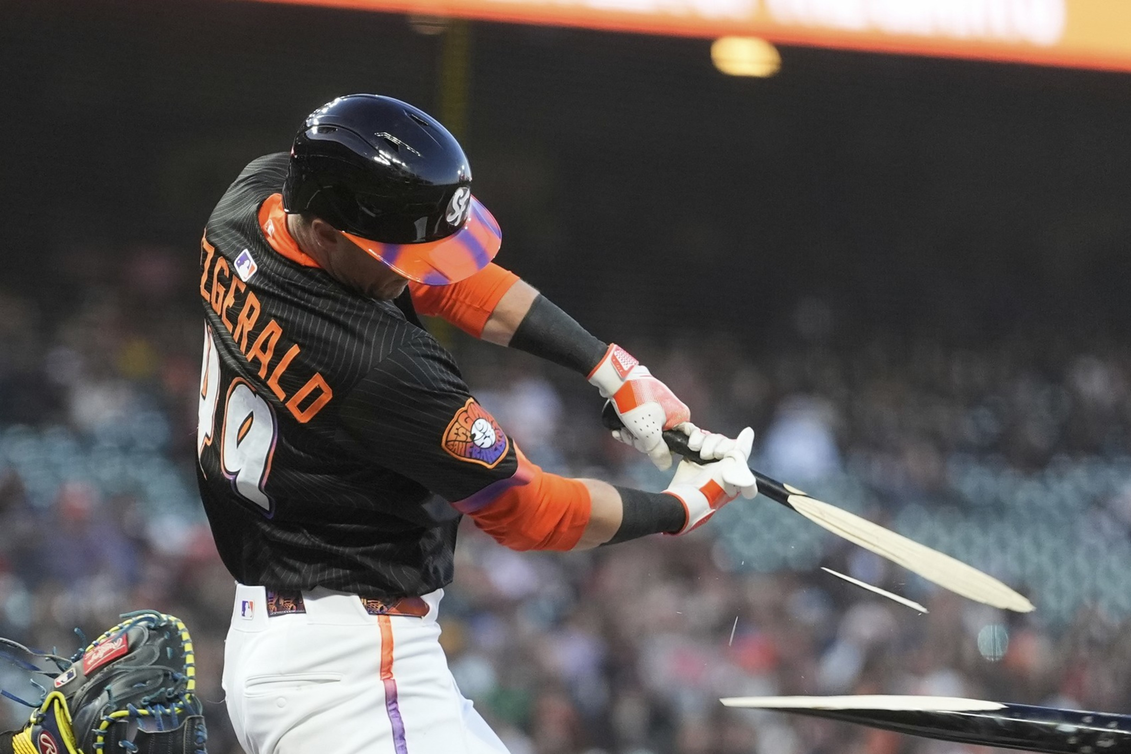 A baseball player in a black jersey swings and breaks his bat as he hits the ball, with the shattered bat pieces flying in opposite directions.