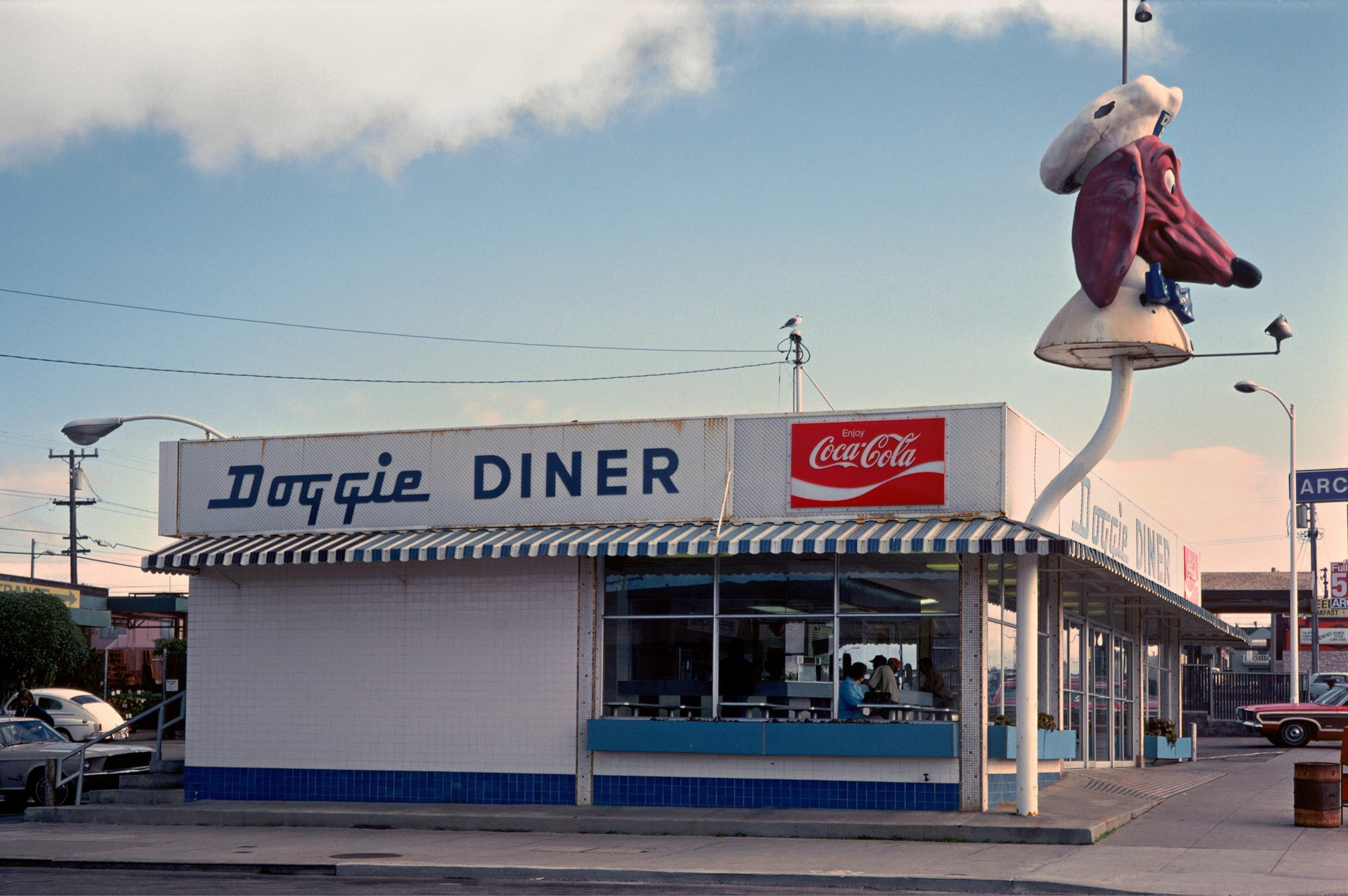 The image shows a retro diner called &quot;Doggie Diner,&quot; featuring a large dachshund head sign wearing a chef's hat above the building. A Coca-Cola sign is visible.