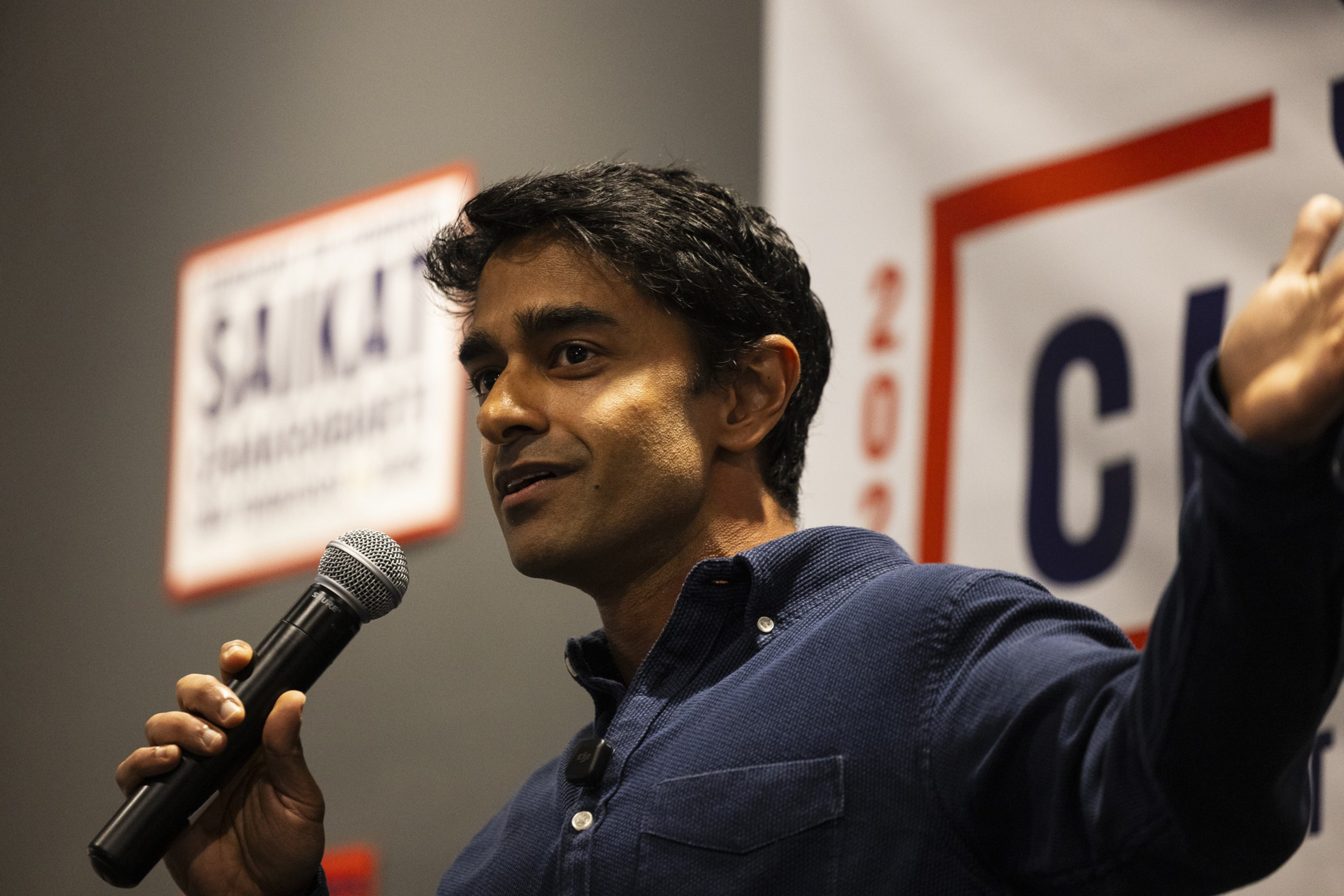 A man in a dark blue shirt speaks into a microphone with one hand, gesturing with the other, standing in front of blurred campaign signs.