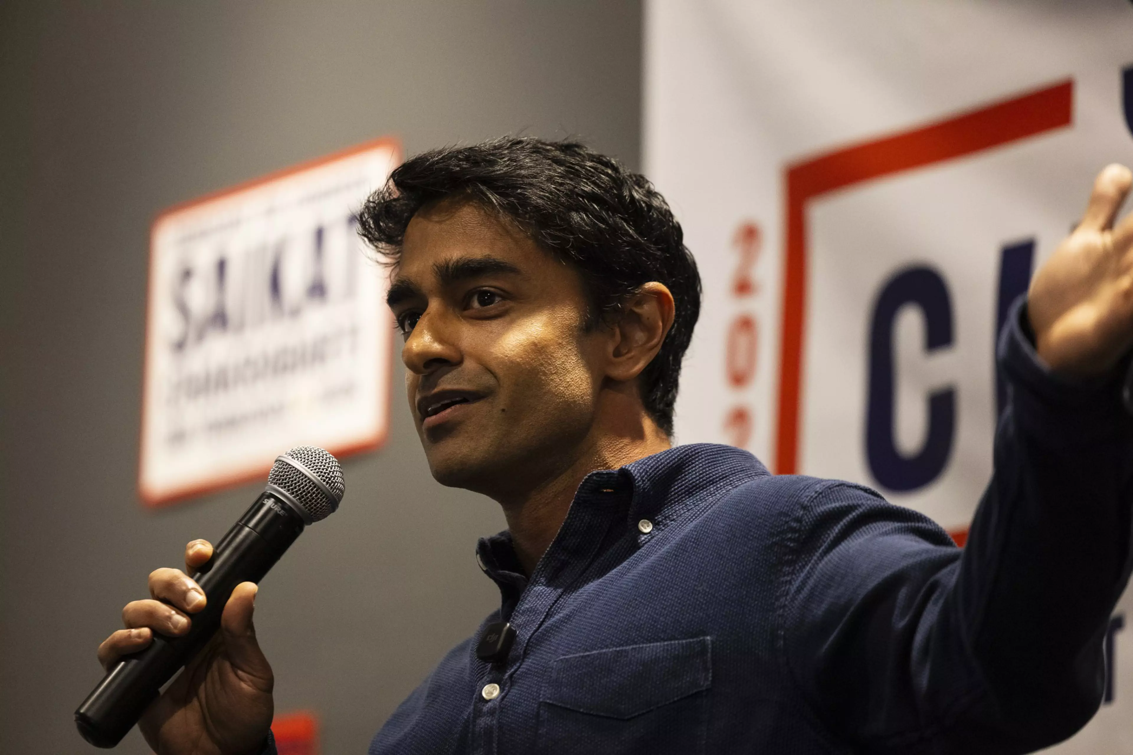 A man in a dark blue shirt speaks into a microphone with one hand, gesturing with the other, standing in front of blurred campaign signs.