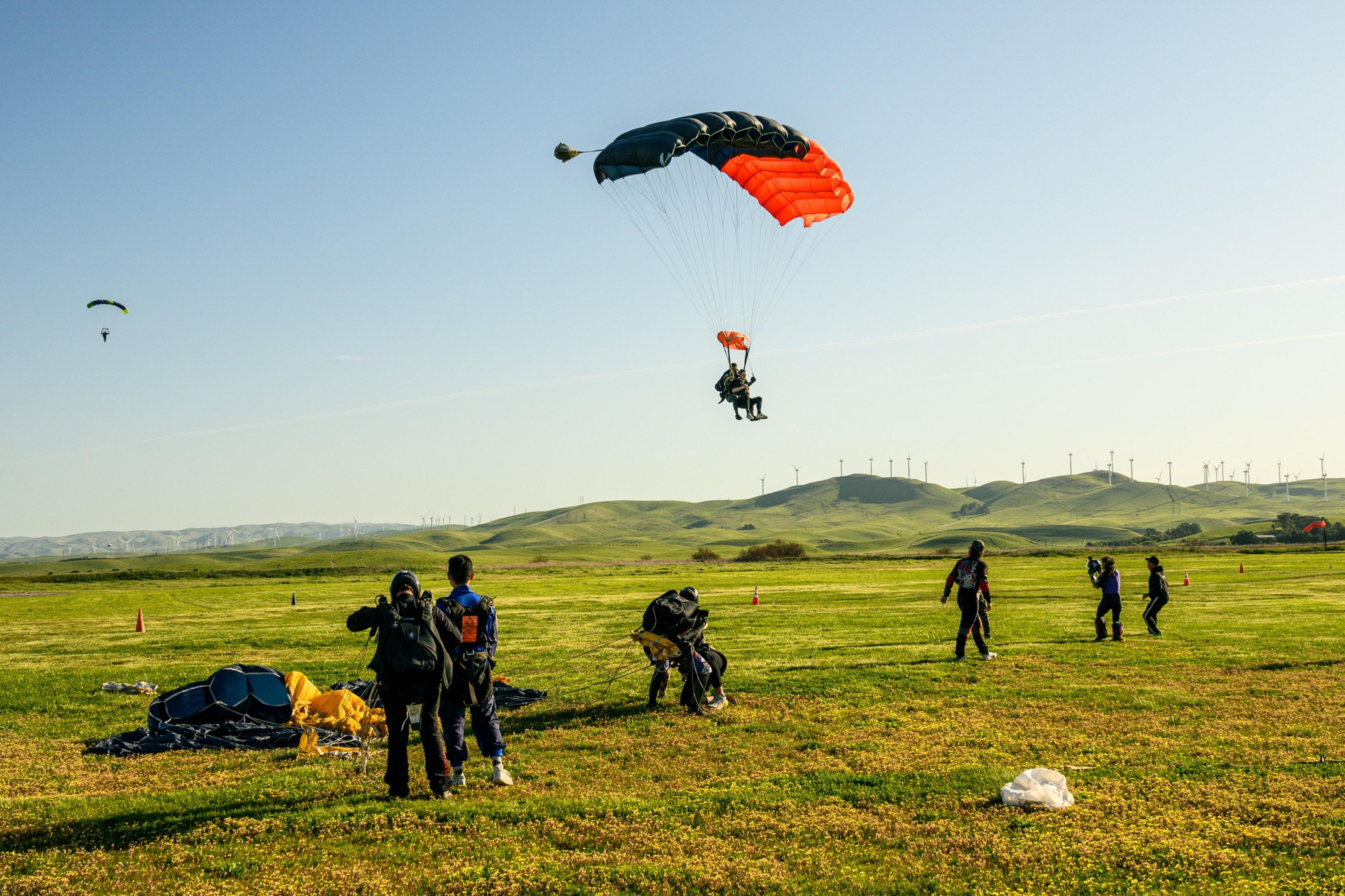 Skydivers land on a grassy field with parachutes open, while others prepare or watch, against a backdrop of green hills and wind turbines.