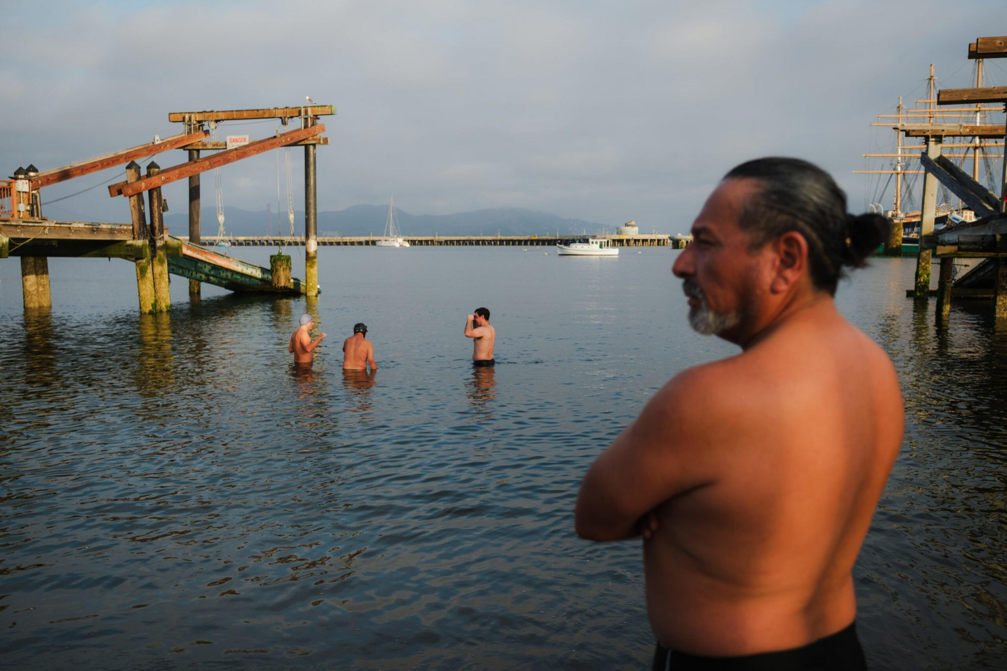 A shirtless man with a ponytail stands with arms crossed near the shore while three other shirtless men wade in the water near a wooden dock.