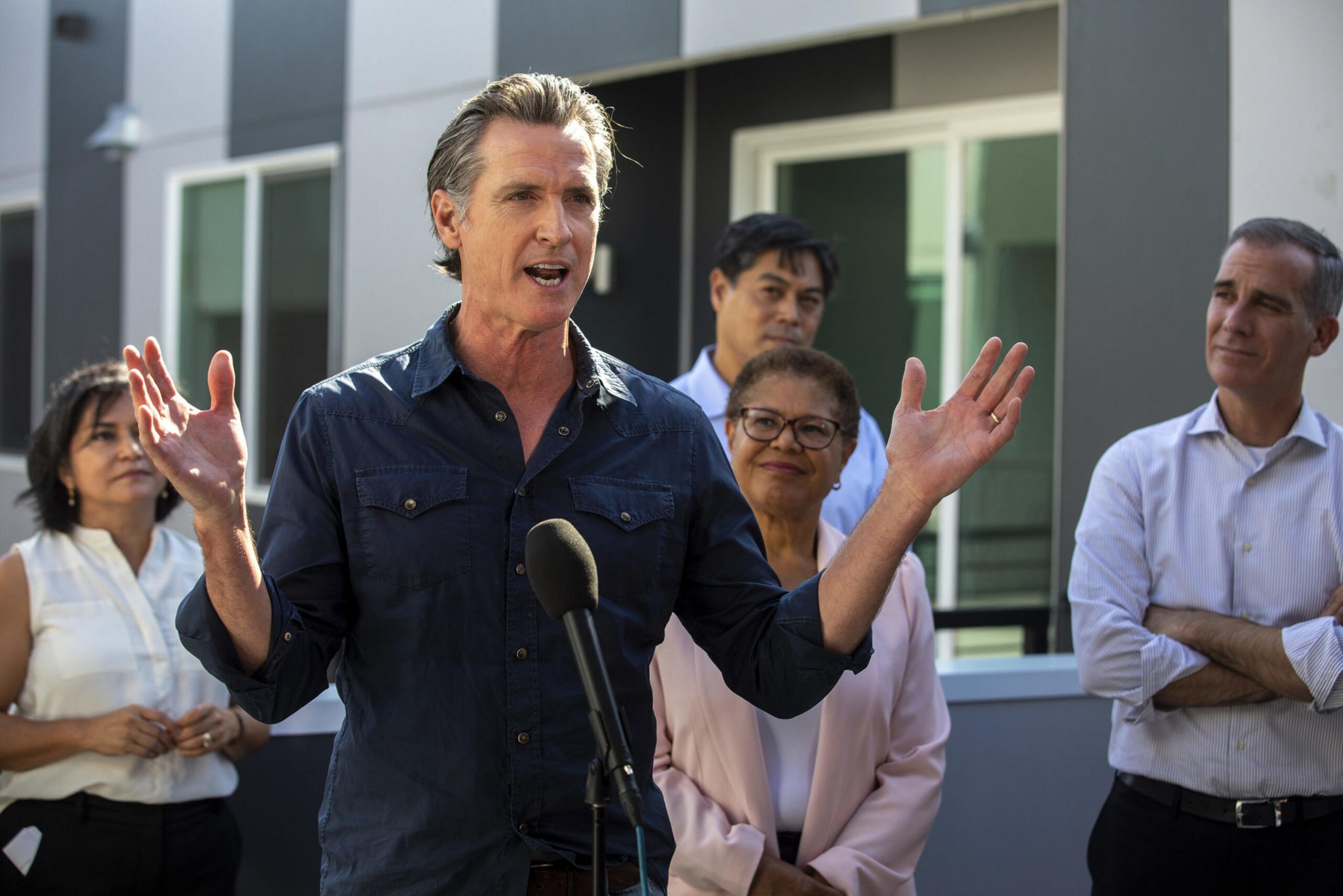 A man speaks passionately at a microphone with four attentive people standing behind him outside a modern building.
