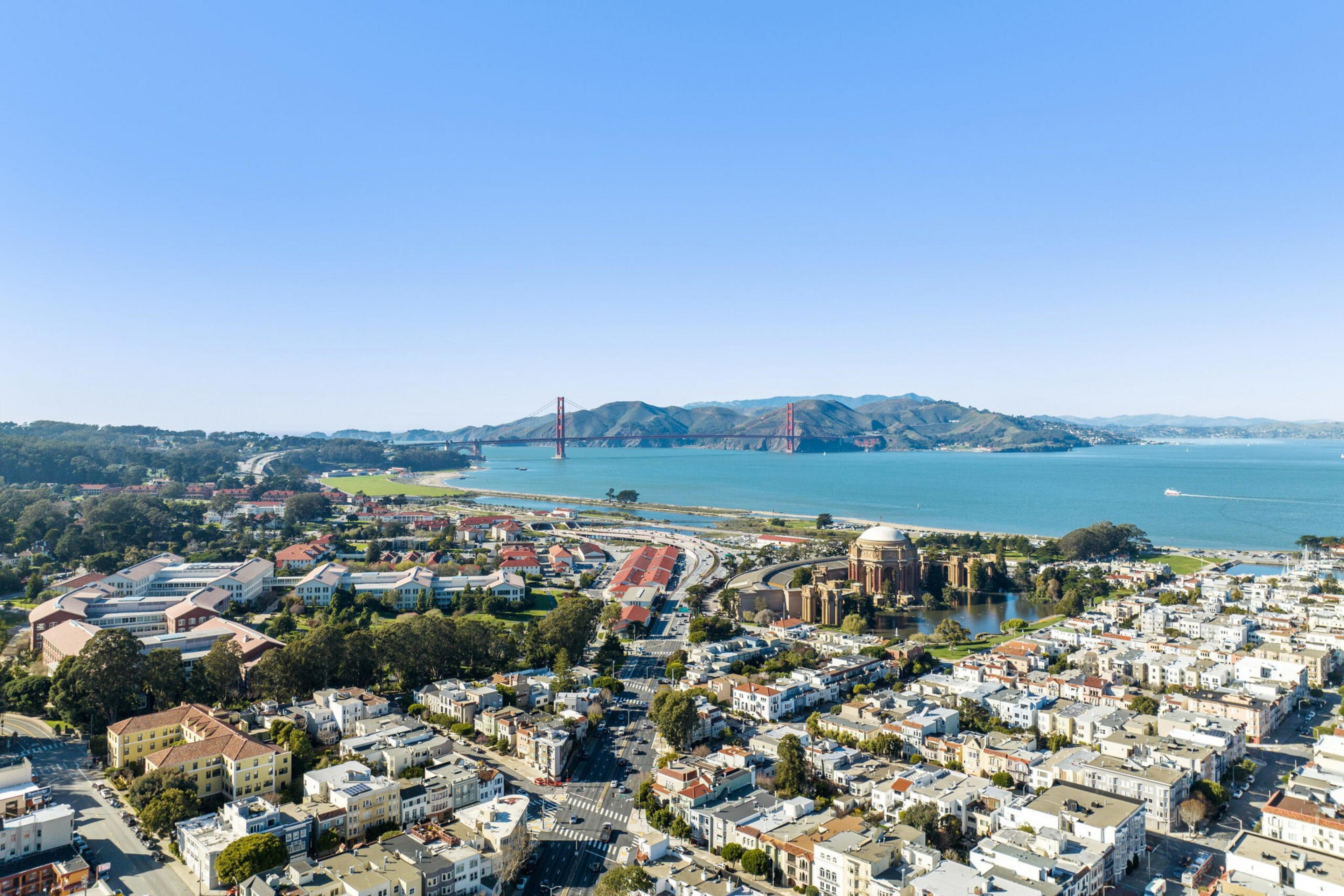 A coastal city with densely packed buildings, a large domed structure by water, a wide bay, green hills, and a red suspension bridge in the distance.